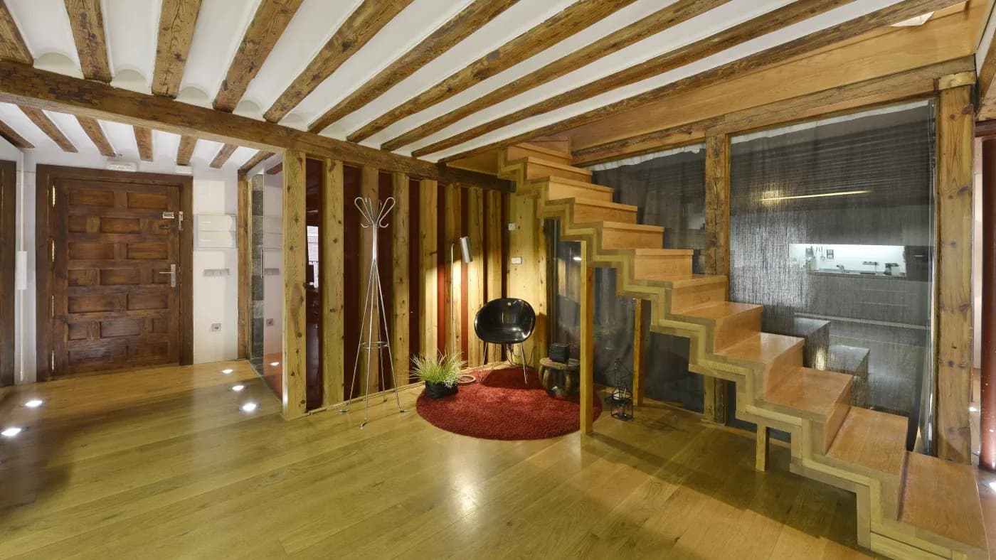 Spacious interior with wooden beams and floor, modern open staircase, a black chair on a red rug, and a plant under warm lighting. Cozy and rustic ambiance.