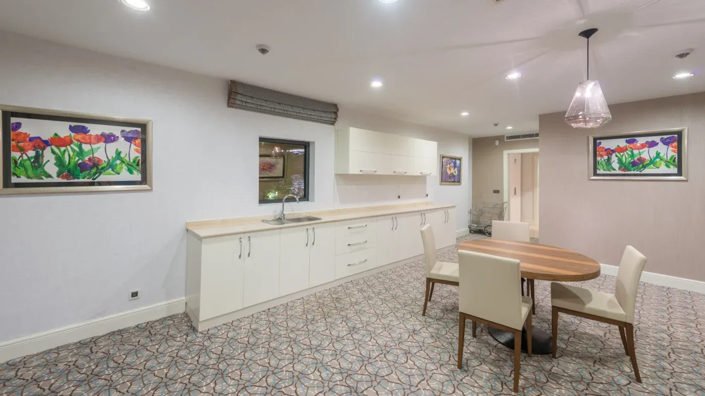 A modern room with white cabinets and a small round table with four white chairs on a patterned carpet. Colorful floral paintings adorn the walls, creating a bright and inviting atmosphere.