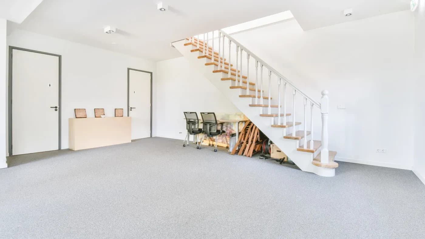 A spacious, bright room with a minimalist design. It features white walls, a carpeted floor, a wooden staircase, a small desk with four awards, and two chairs.