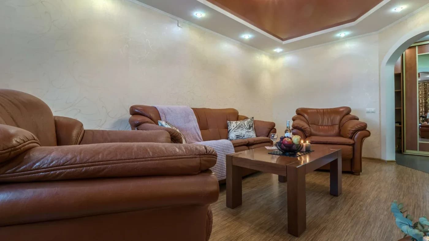 A cozy living room featuring brown leather sofas, a wooden coffee table with candles and decor, warm lighting, and a textured beige wall.