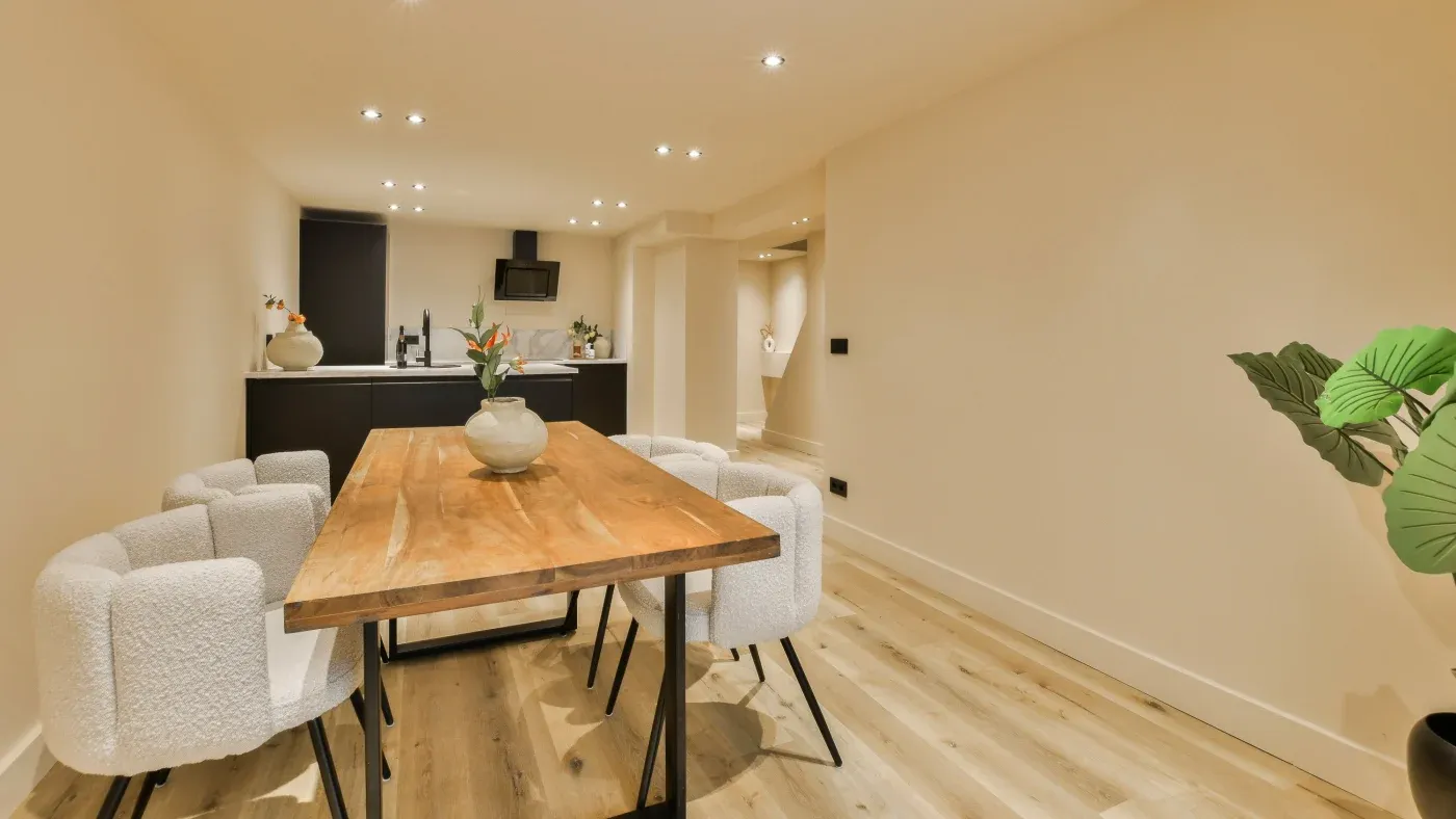 Modern dining area with a wooden table, six white cushioned chairs, and a minimalist kitchen in the background. Bright, cozy, and inviting atmosphere.