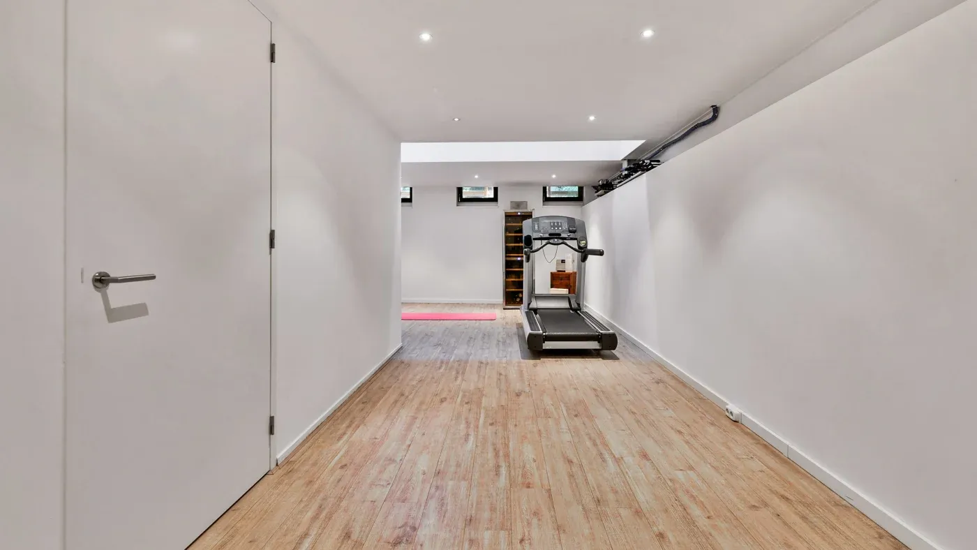 A minimalist basement gym with a treadmill on a wooden floor, recessed lighting, and small windows. The open space conveys a clean, simple atmosphere.