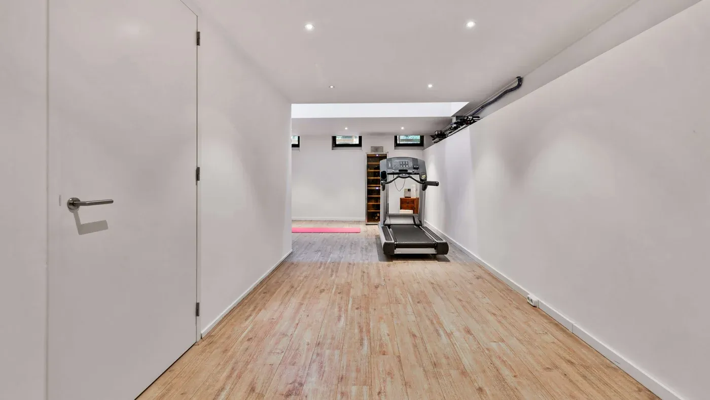 A minimalist home gym with a treadmill at the end of a narrow, wood-floored room. White walls and recessed lighting create a clean, modern feel.