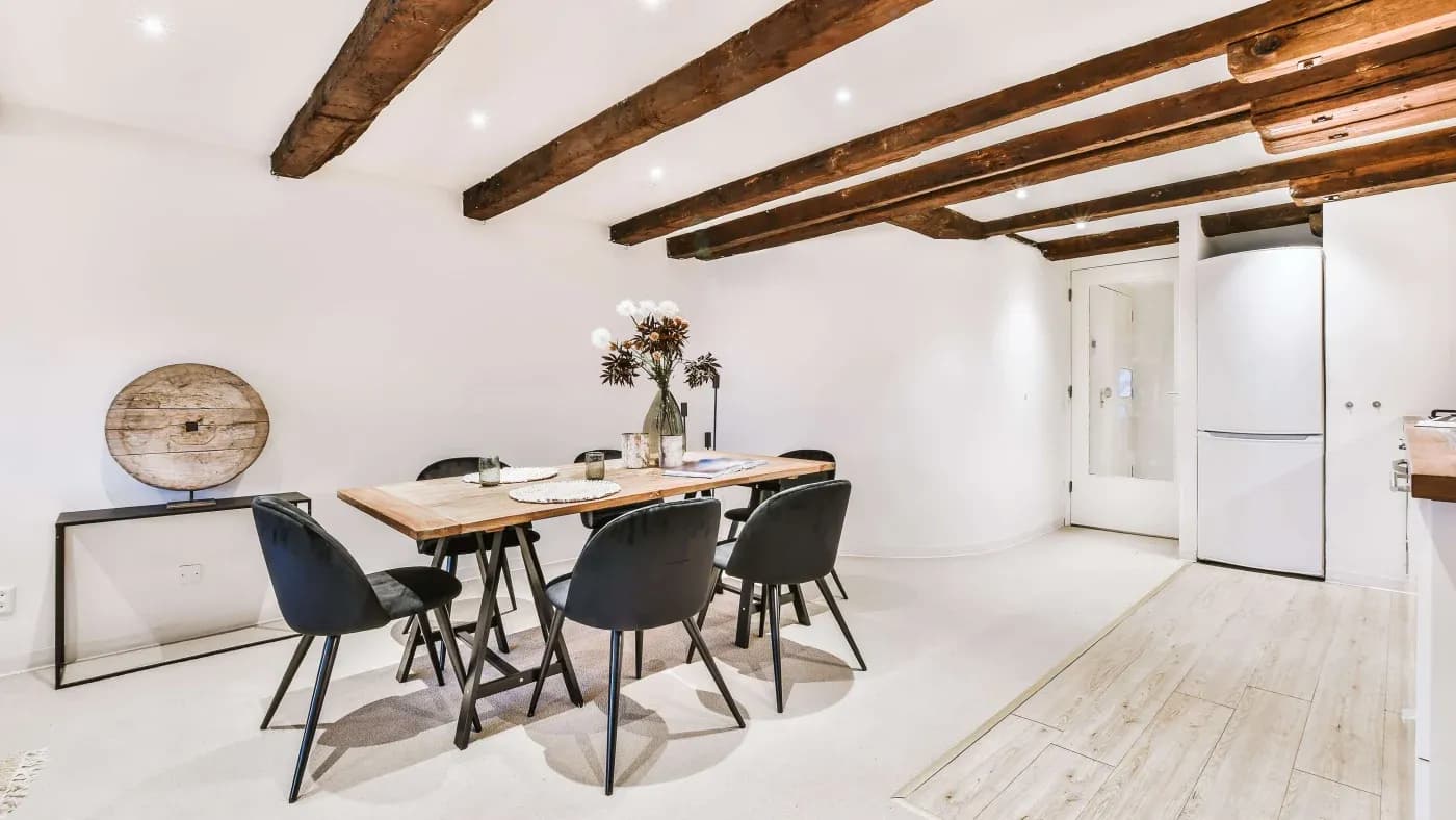 A cozy dining room with wooden ceiling beams, a wooden table set with six black chairs, a vase of flowers, recessed lighting, and a neutral color palette.