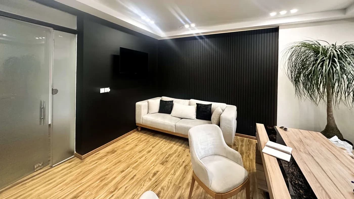 A modern office space with a light wood floor, a white three-seater sofa with black cushions, a flat-screen TV on a black wall, and a sleek wooden desk.