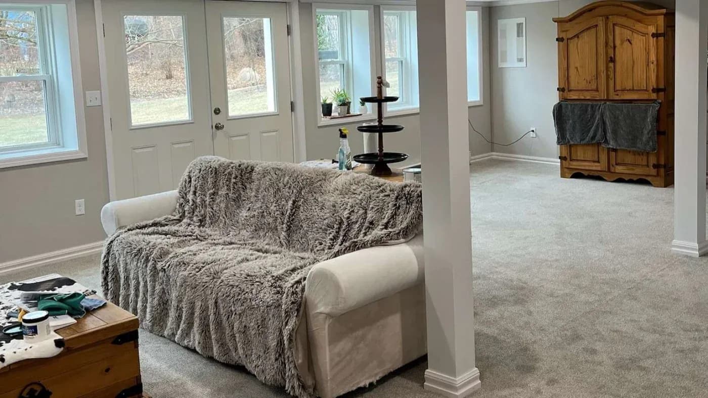 A cozy basement with a fluffy gray throw on a white sofa, colorful game controllers on a wooden chest, and a wooden cabinet. Soft light fills the room.