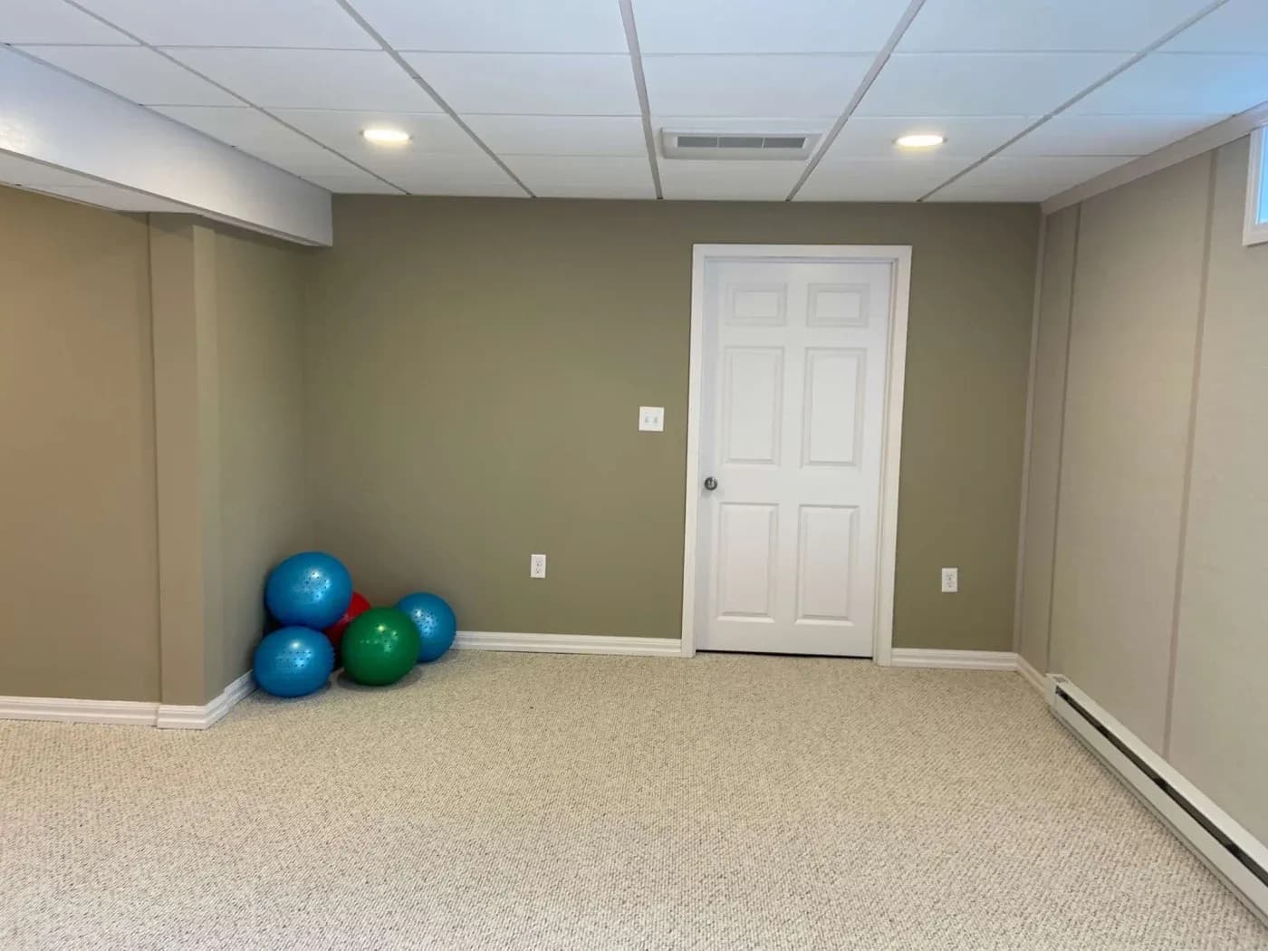 Finished basement with olive-green walls, white dropped ceiling with recessed lighting, beige carpet, white paneled door, and three exercise balls stacked in corner
