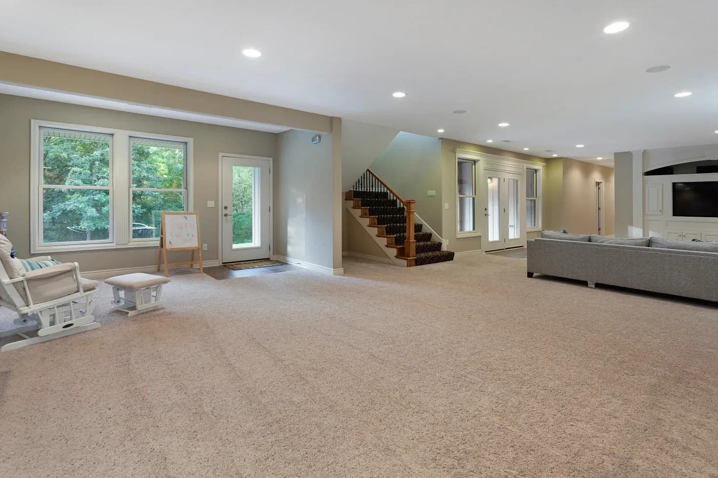 Spacious basement with beige carpet, large windows, a staircase, and a gray sectional sofa. Bright natural light and a calm, inviting atmosphere.