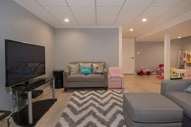 A cozy basement living room with gray walls, a plush sofa, patterned rug, and a TV. Toys are scattered, adding a playful touch. Soft lighting creates a warm, inviting atmosphere.