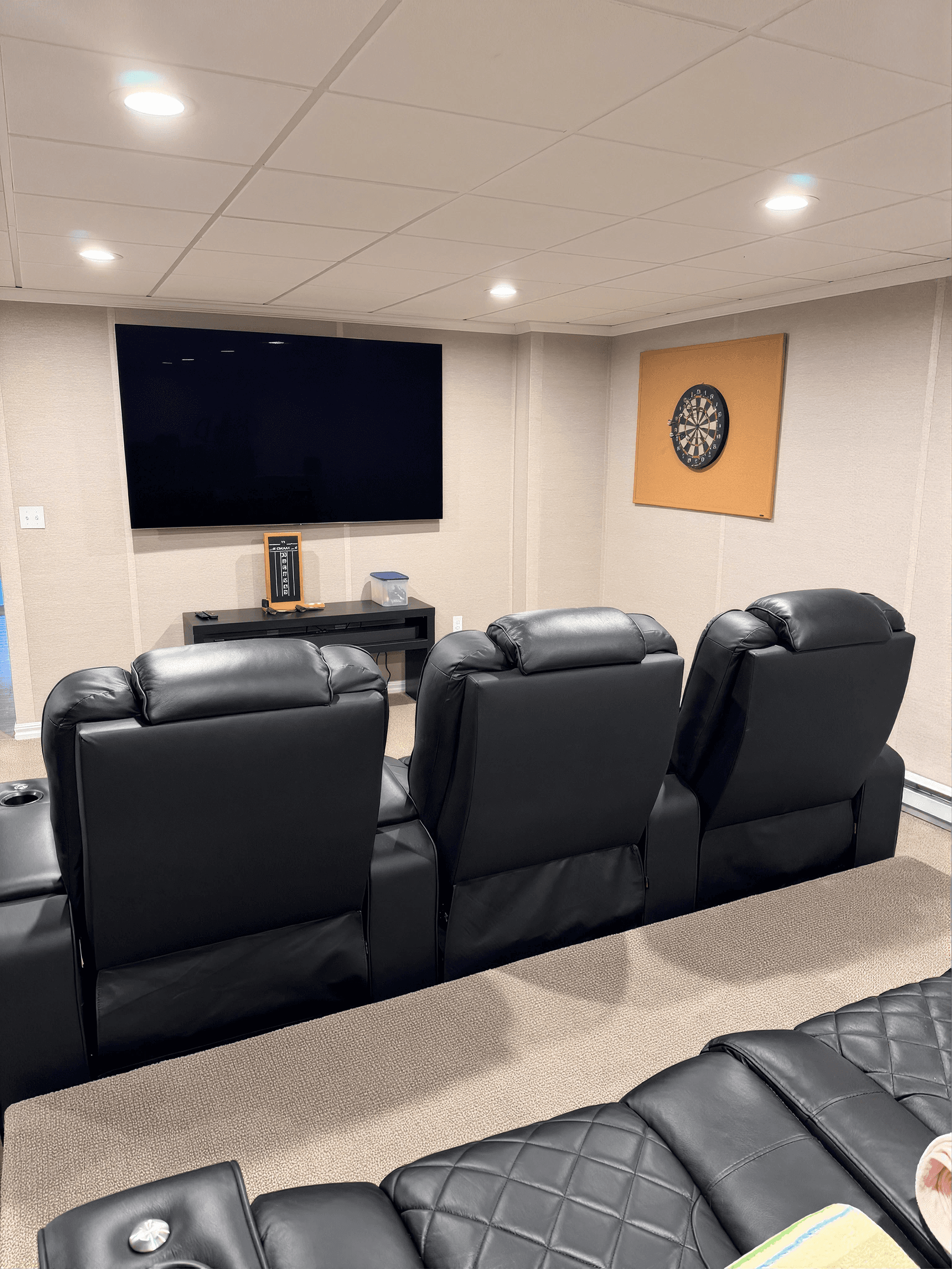 A cozy home theater with three black leather recliner seats facing a large flat-screen TV. A dartboard decorates the beige wall, creating a relaxed atmosphere.