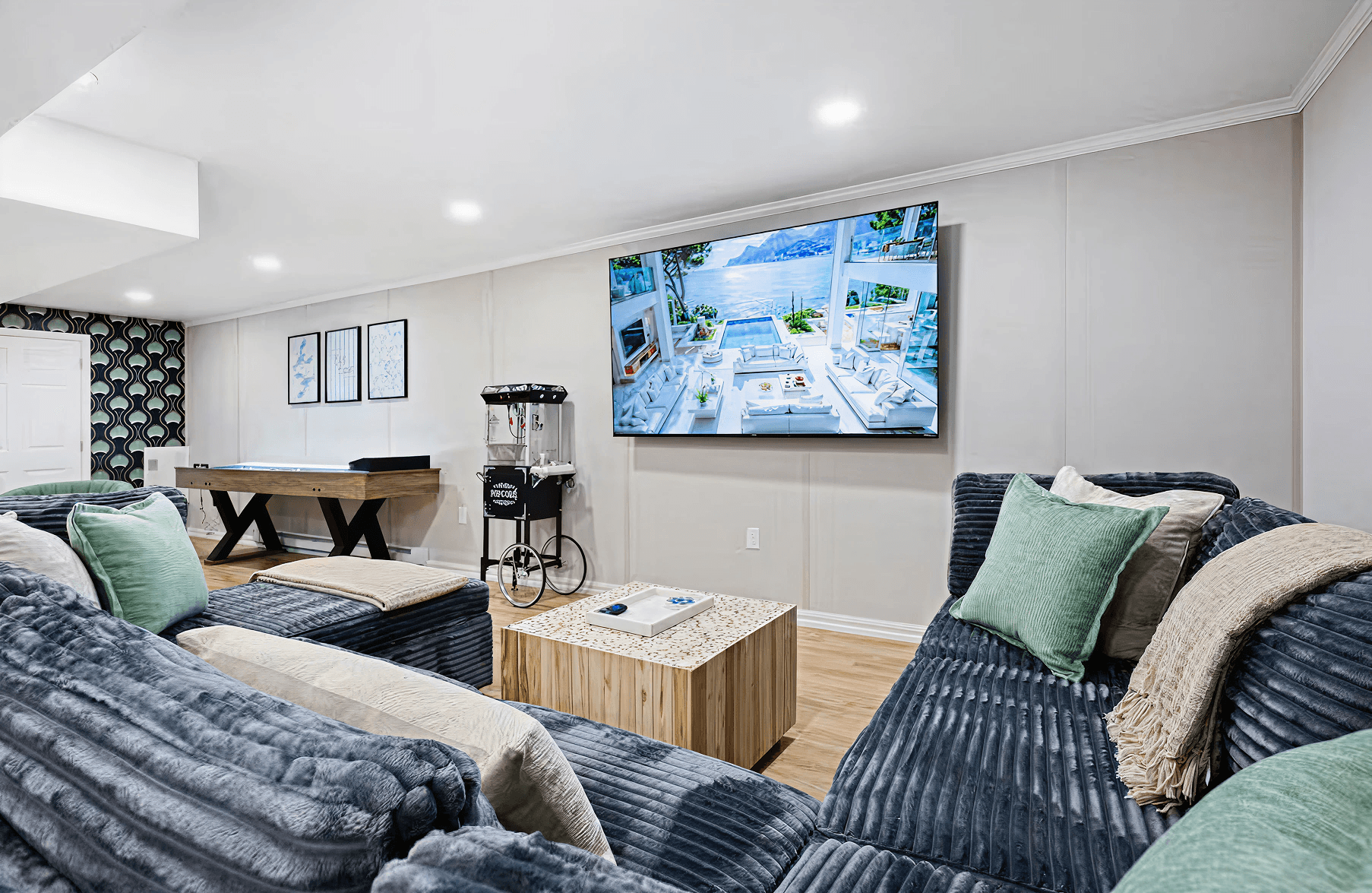 Stylish basement entertainment room with cozy blue couches, plush pillows, a large wall-mounted TV, air hockey table, and popcorn machine.