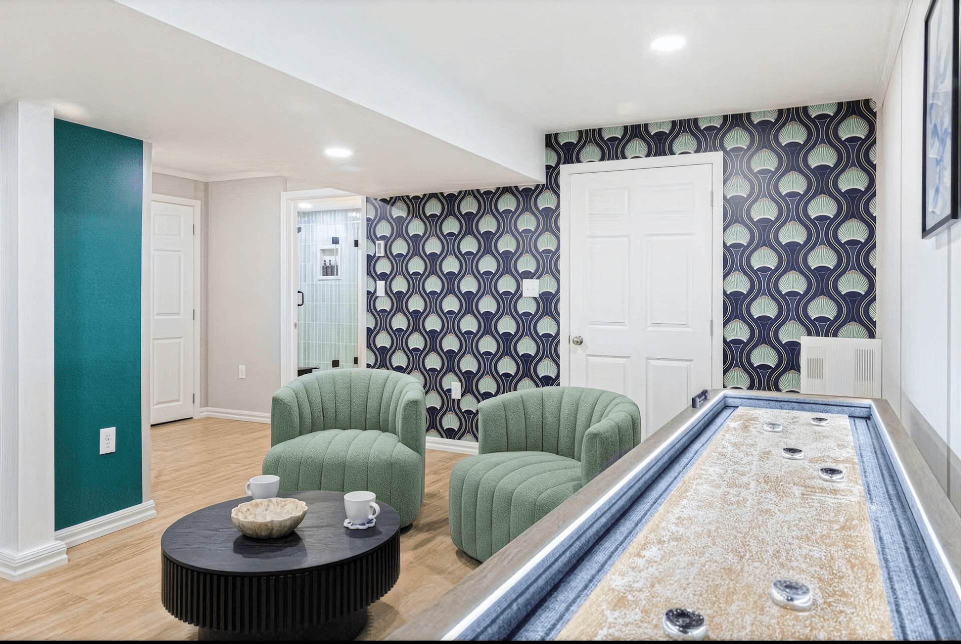 Stylish living area with retro-patterned wallpaper, two green chairs, and a black round table with cups. A shuffleboard table adds a playful touch.