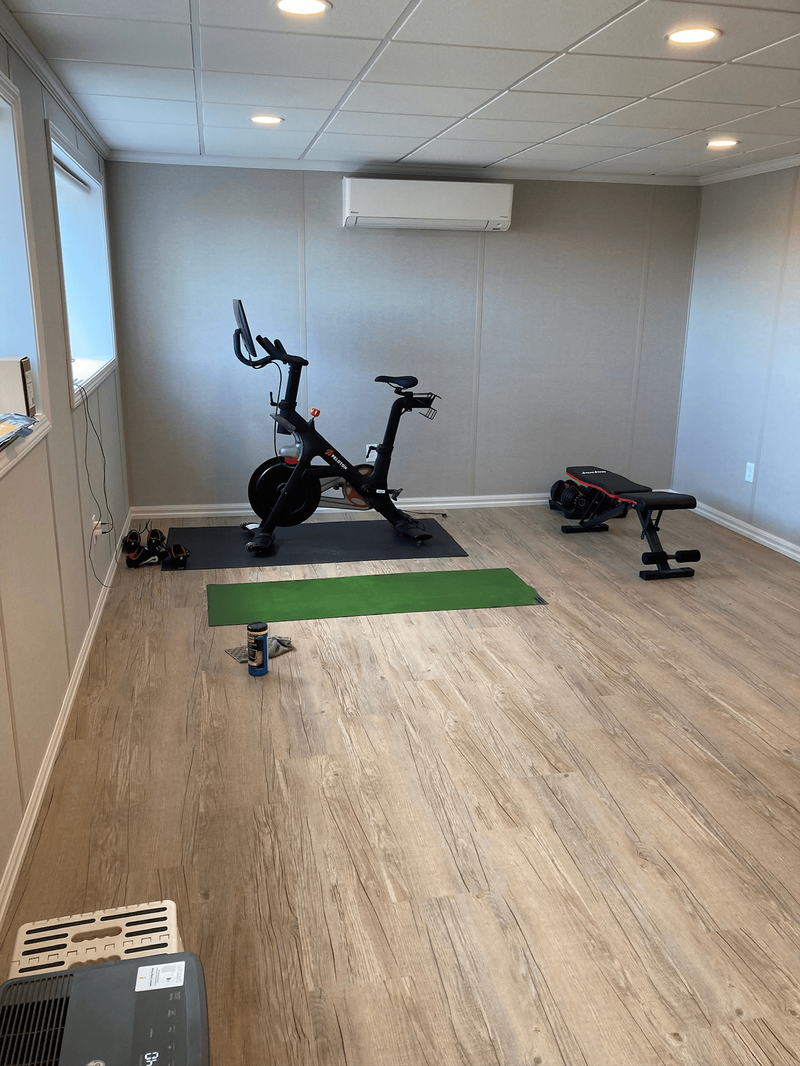 Home gym with an exercise bike, weight bench, and green yoga mat on wood flooring. Bright lighting and a minimal, clean atmosphere.