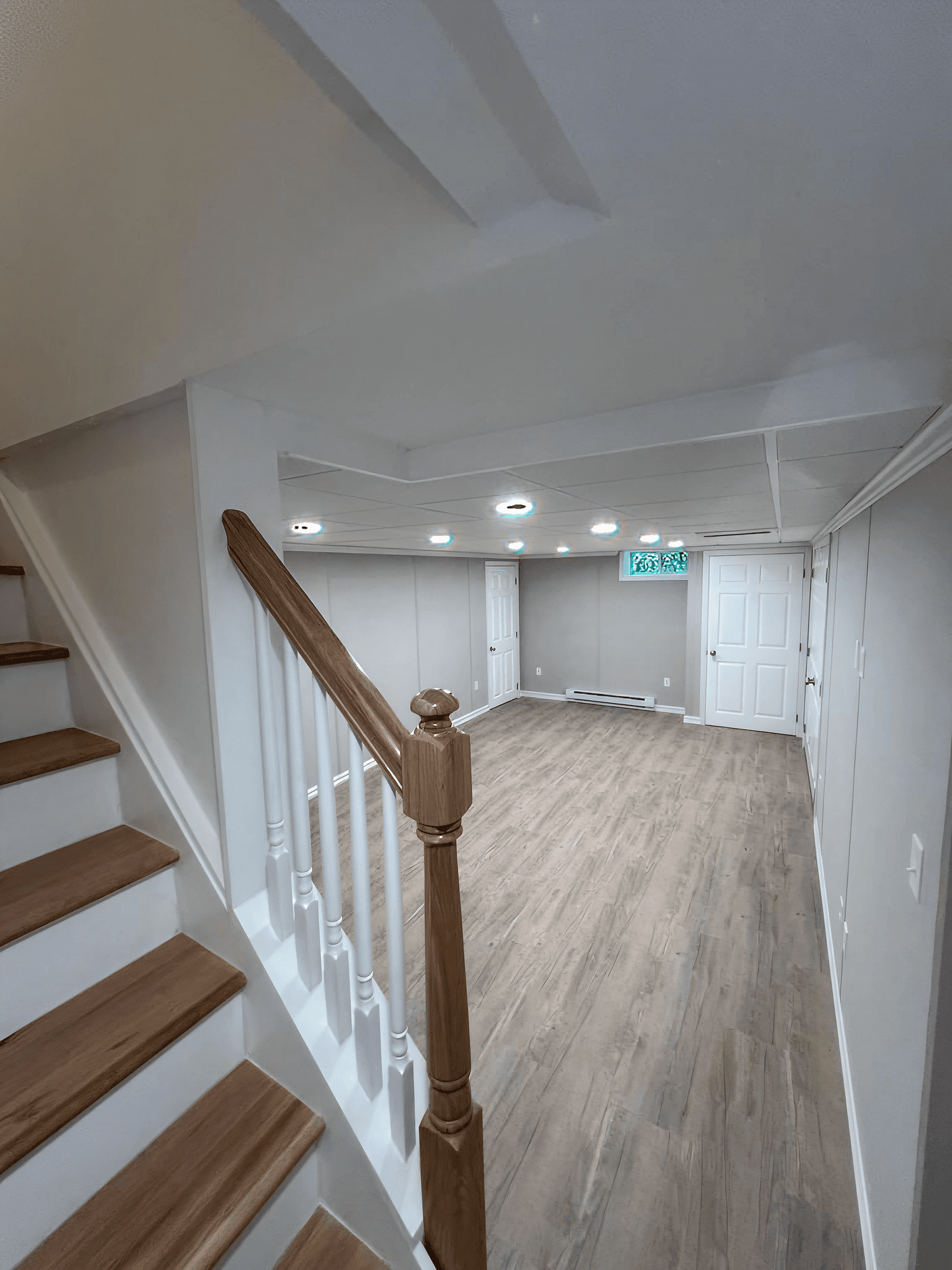 A well-lit, newly renovated basement with light wood flooring, gray walls, and recessed lighting. A wooden staircase leads down into the open space.