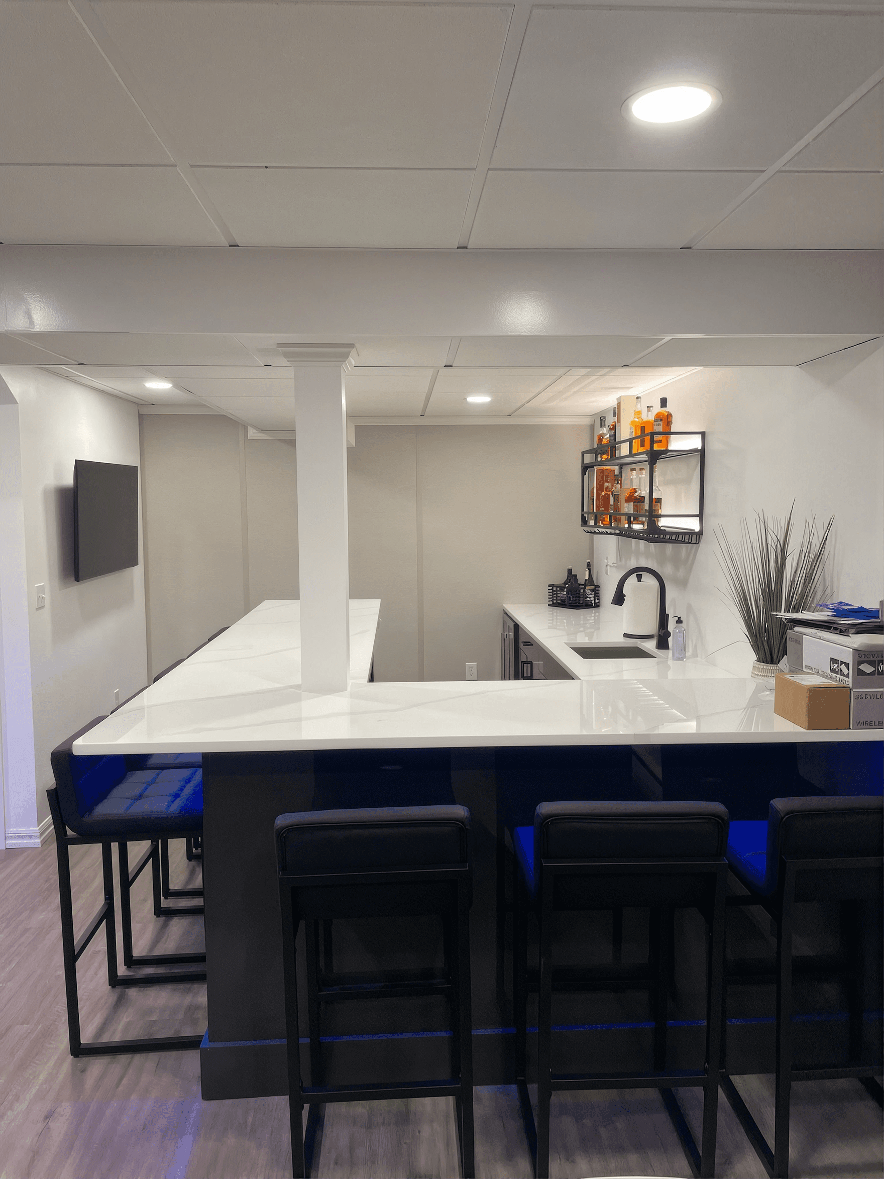 Modern home bar with white countertops, black stools, and blue cushions. Wall-mounted shelves hold bottles, creating a sleek, inviting atmosphere.