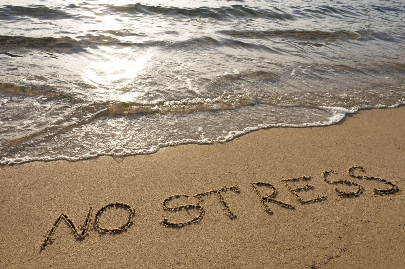 The image shows the words "NO STRESS" written in the sand on a beach, with gentle waves lapping at the shore. The scene conveys relaxation and calm.
