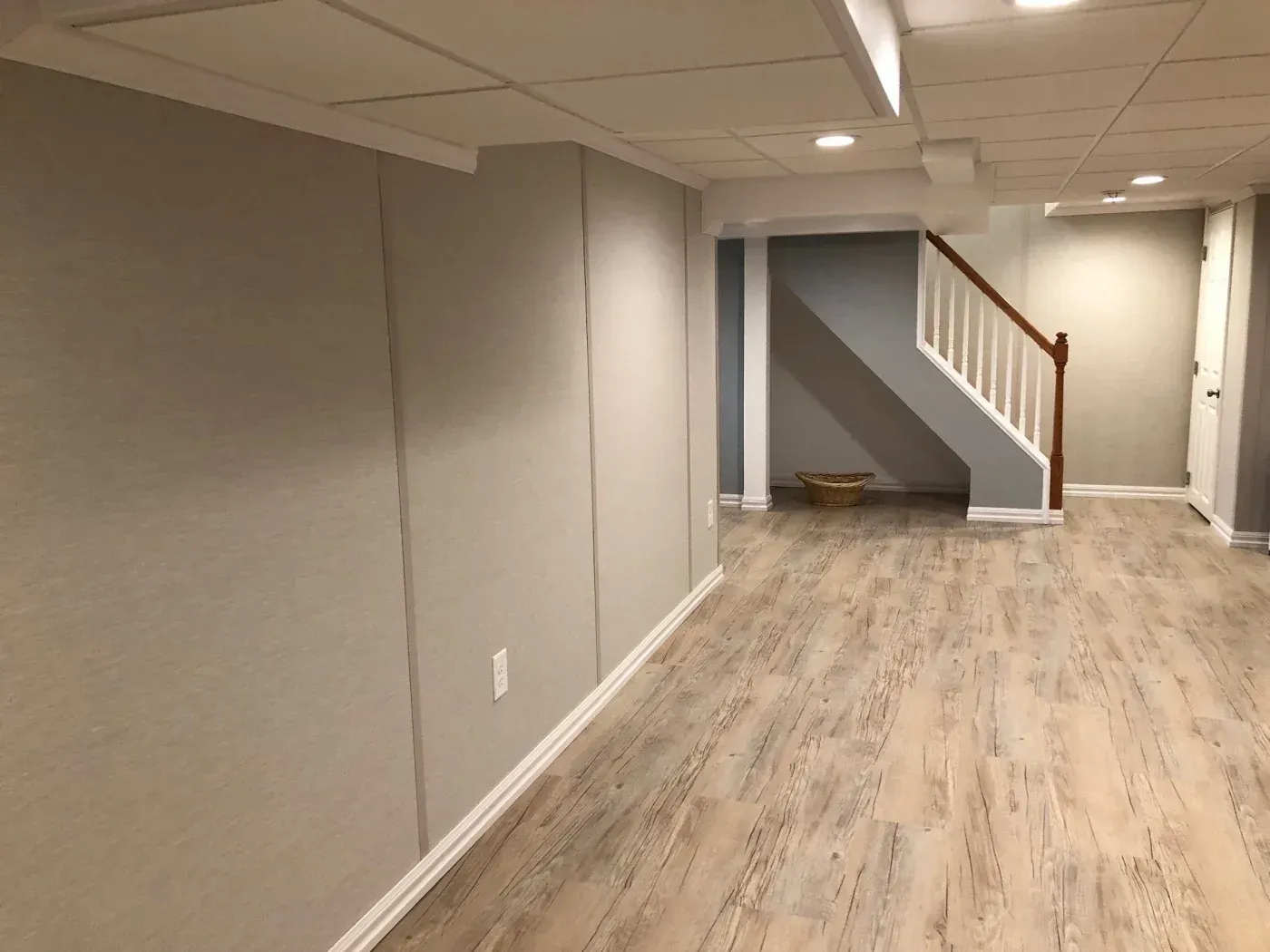 A modern basement with light wood flooring, neutral walls, and recessed lighting. An open staircase with white railings adds a cozy, minimal feel.