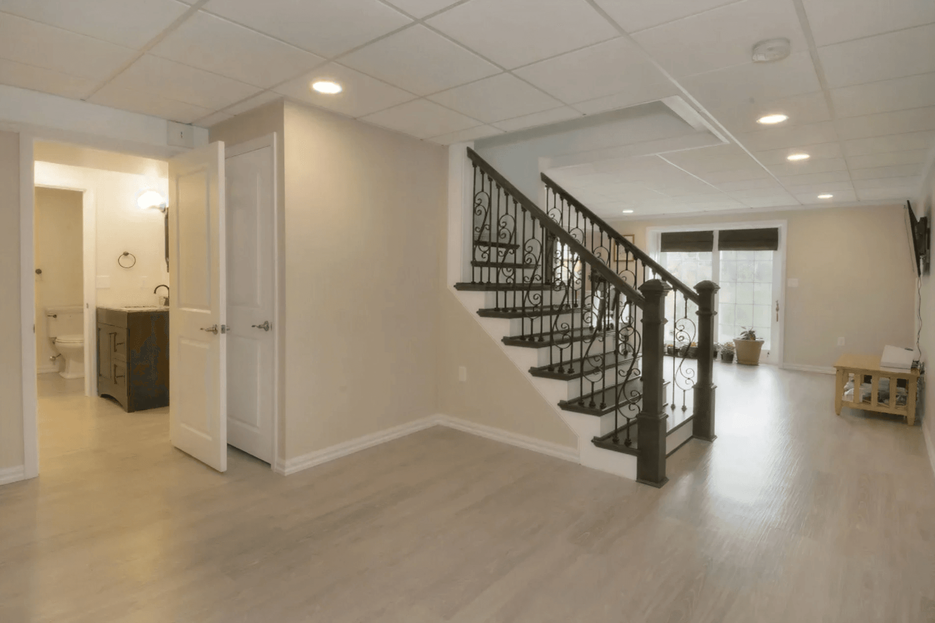 A modern basement with light wood flooring, white walls, and a ceiling with recessed lights. A staircase with ornate railings leads upstairs. A white door opens to a small bathroom. Large windows brighten the space, with a small table and potted plant near the entrance, creating an inviting atmosphere.