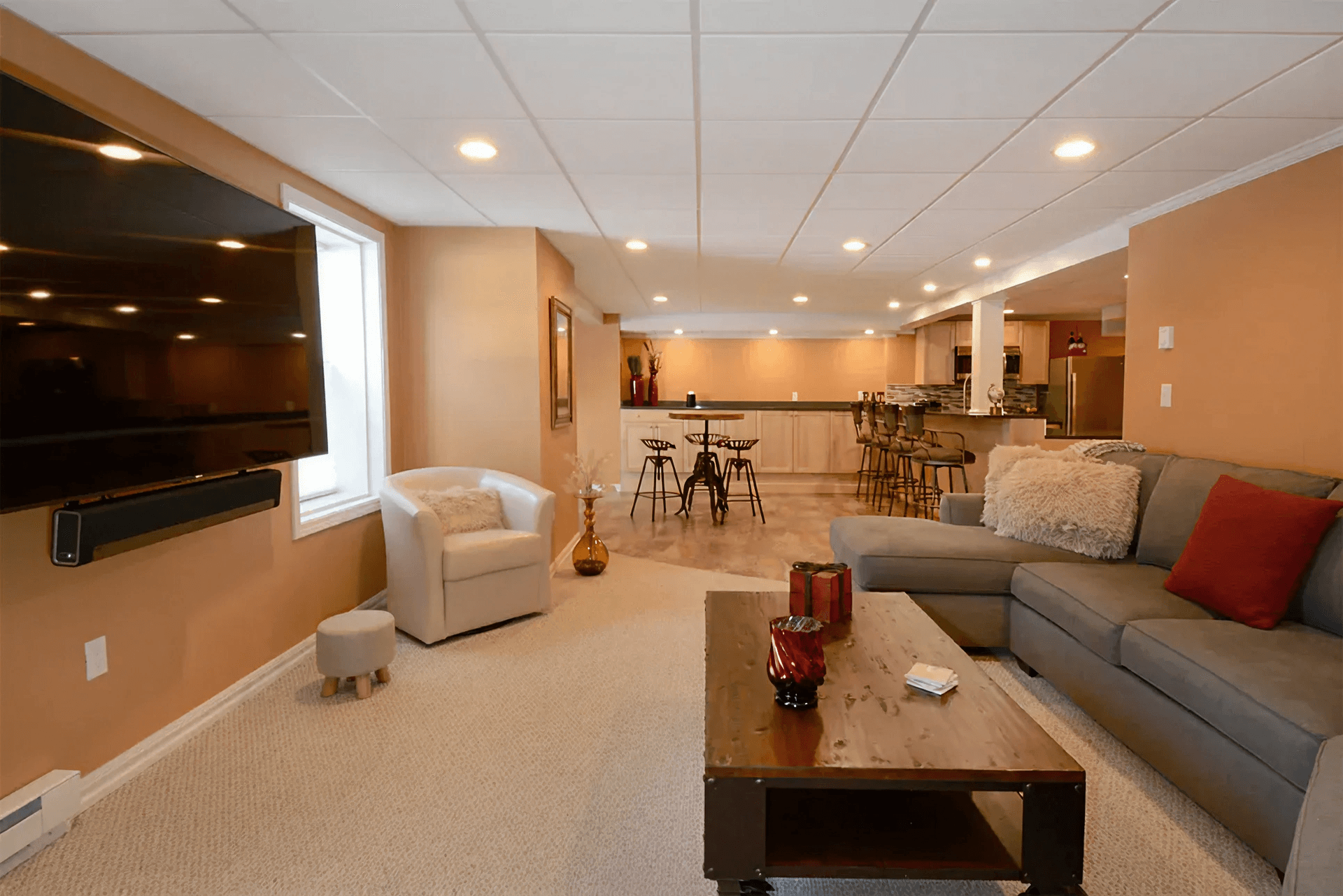 Spacious basement with beige walls, gray sectional sofa, and wall-mounted TV. Features a bar area with stools and a cozy atmosphere.
