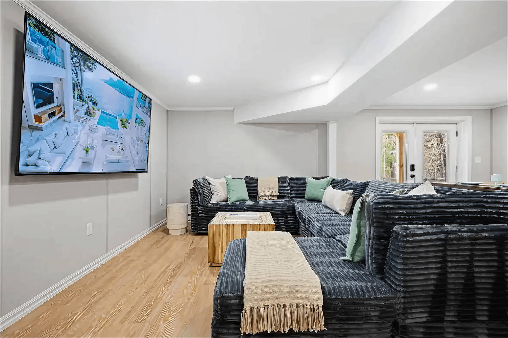 Spacious living room with a large sectional sofa adorned with green and beige cushions and a throw, facing a wall-mounted flat-screen TV. Neutral tones and soft lighting create a cozy atmosphere.