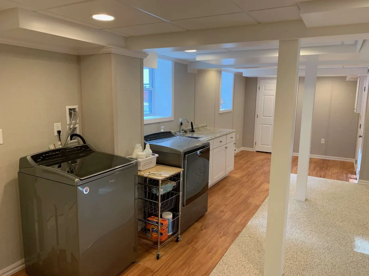 Modern laundry room with washer and dryer, gray countertops, white cabinets, and wooden floors. Bright lighting and neutral tones create a clean, organized atmosphere.