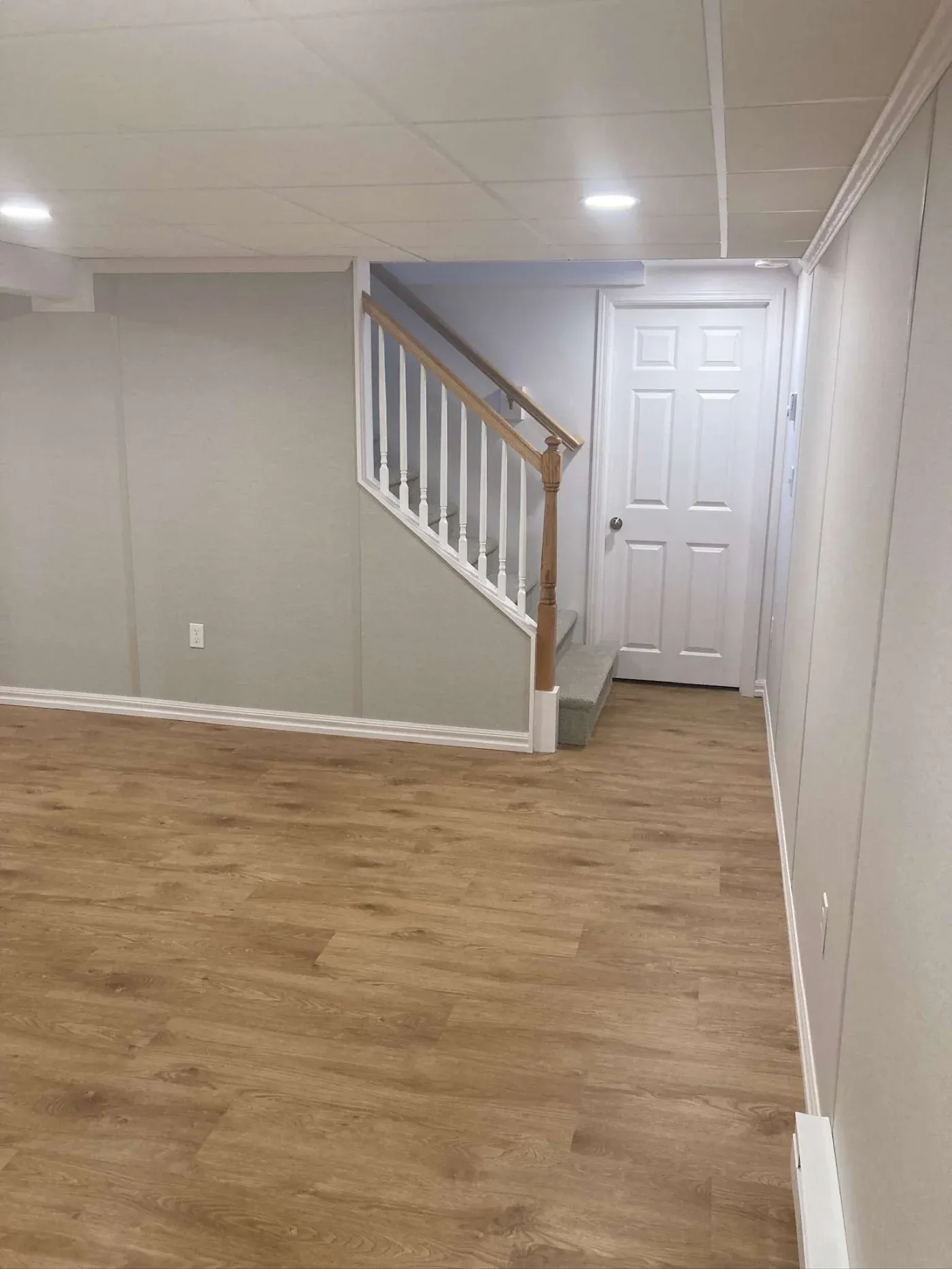 A bright, empty basement with light beige walls, wooden flooring, recessed ceiling lights, and a staircase with a wooden handrail leading up to a white door.
