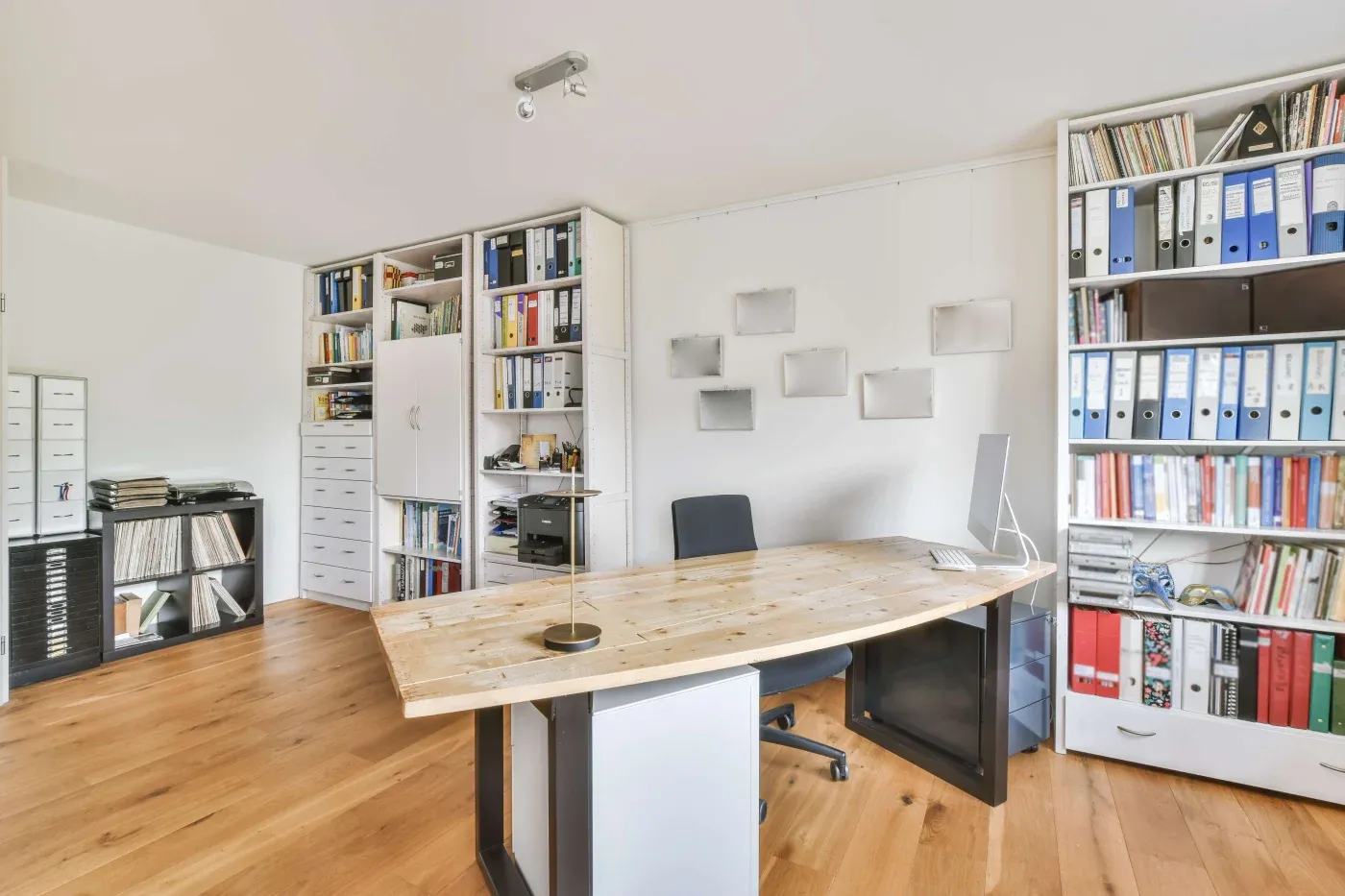 A modern office with a wooden desk, black chair, computer, lamp, and bookshelves filled with binders and books. The space is organized and well-lit.