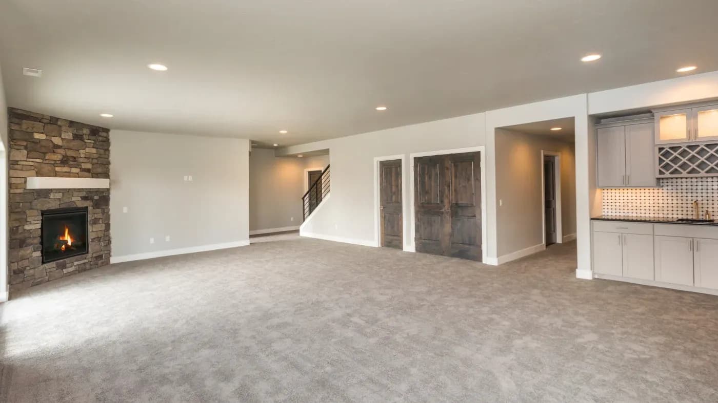Spacious, empty living room with beige carpet, stone fireplace, recessed lighting, and a wet bar with grey cabinets. Neutral tones convey a cozy ambiance.
