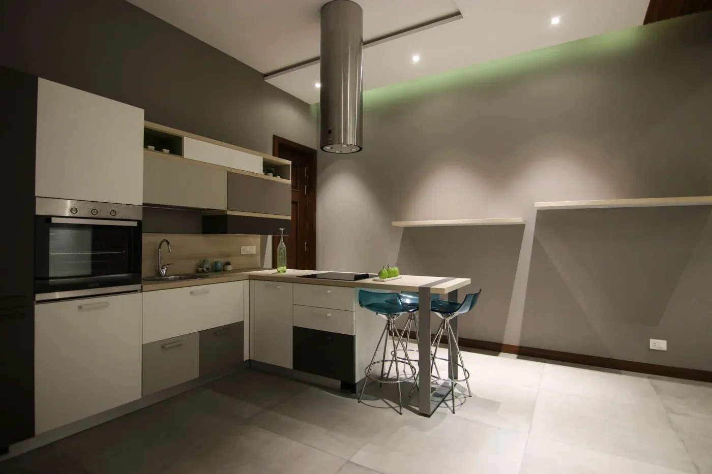 Modern kitchen with sleek grey and white cabinets, built-in oven, and a minimalist island. Two blue stools add color; soft lighting creates a cozy feel.