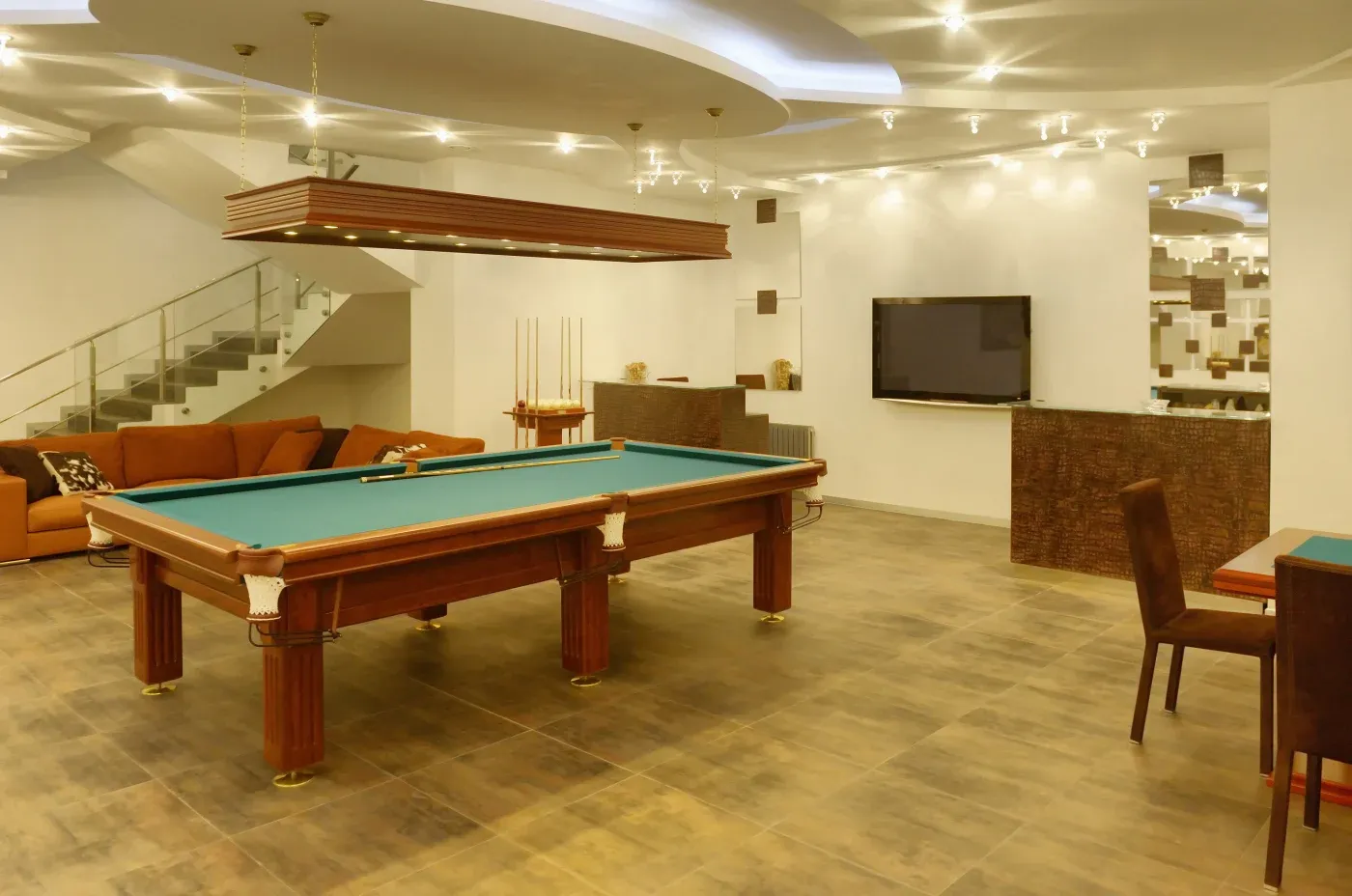 Luxurious recreation room with a central pool table, modern lighting, brown seating area, wall-mounted TV, and stylish decor. Calm, inviting atmosphere.