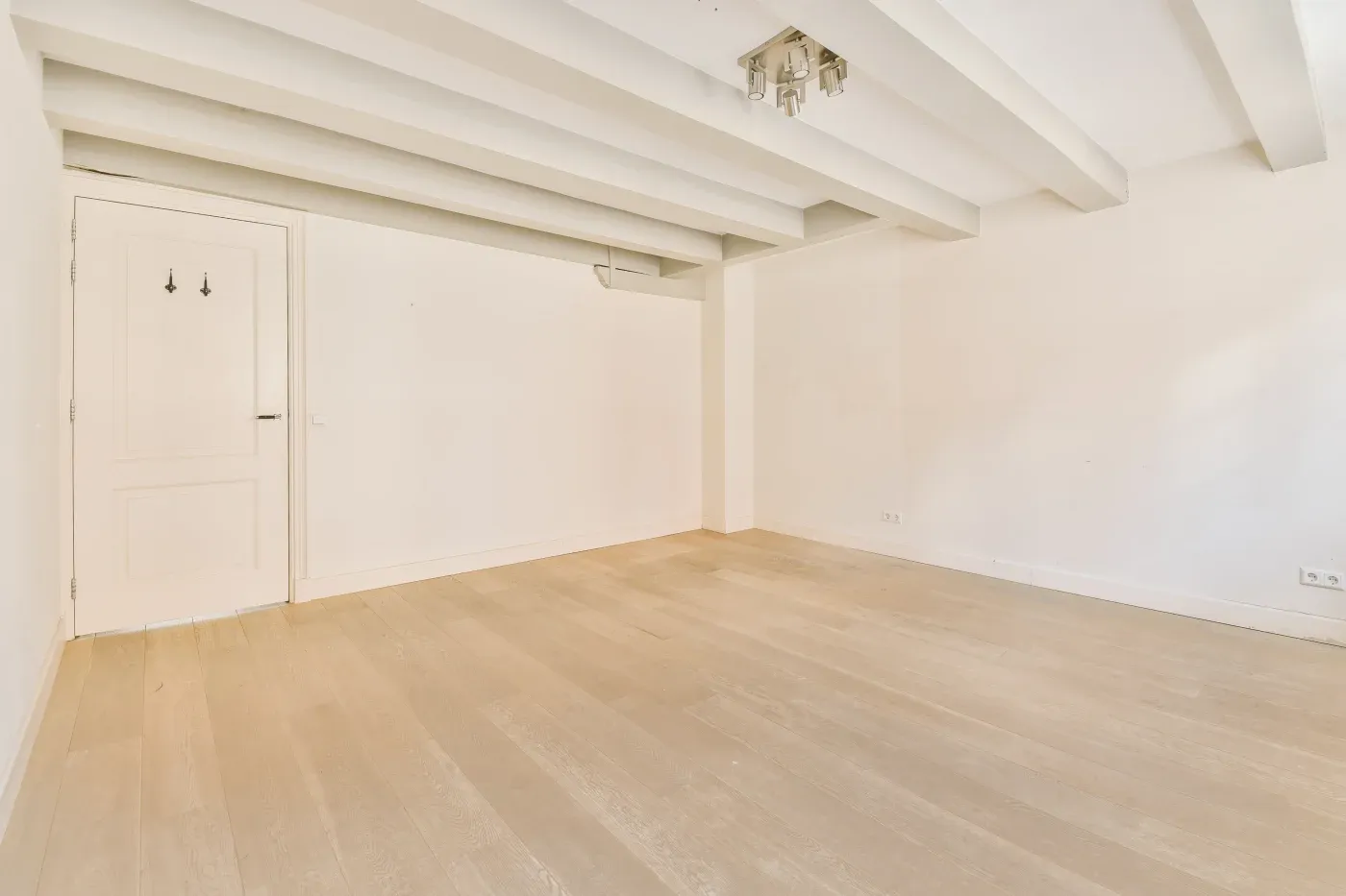 A spacious empty room with light wooden flooring, white walls, and exposed ceiling beams. A closed white door is on the left, and sunlight filters in softly.