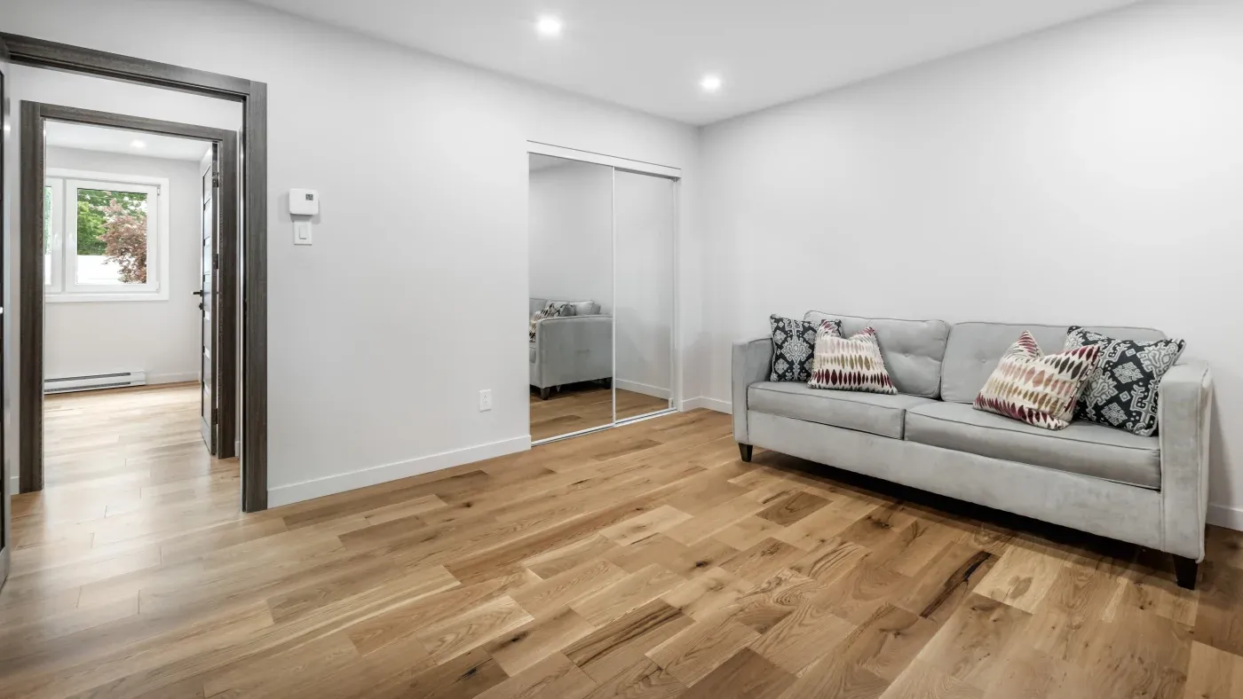 A modern living room with light wood flooring, a gray sofa with patterned cushions, and mirrored closet doors. The room is bright and minimalistic.