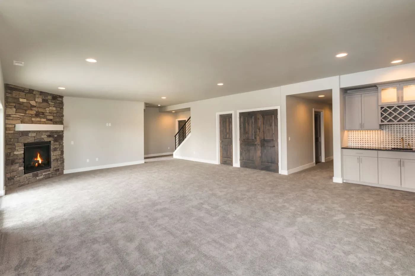 Spacious basement with beige carpet, stone fireplace, wooden doors, and a kitchenette with cabinets. Warm lighting creates a cozy atmosphere.