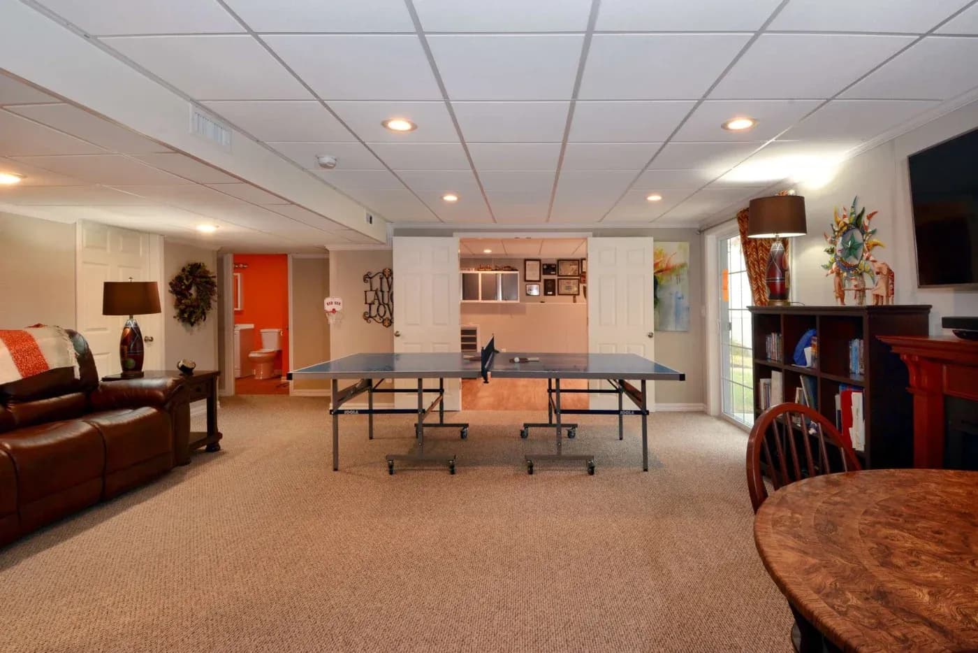 Spacious basement with a ping-pong table at the center, leather sofa, and small dining table. Cozy, inviting ambiance with warm lighting.