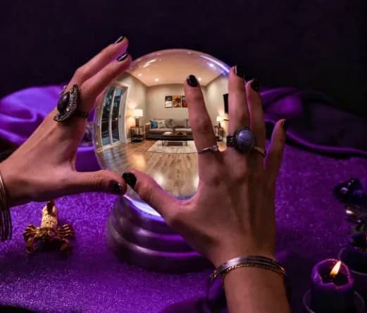 Hands with dark nail polish and rings hold a crystal ball, reflecting a cozy living room. The scene is mystical, with a purple cloth and lit candles.