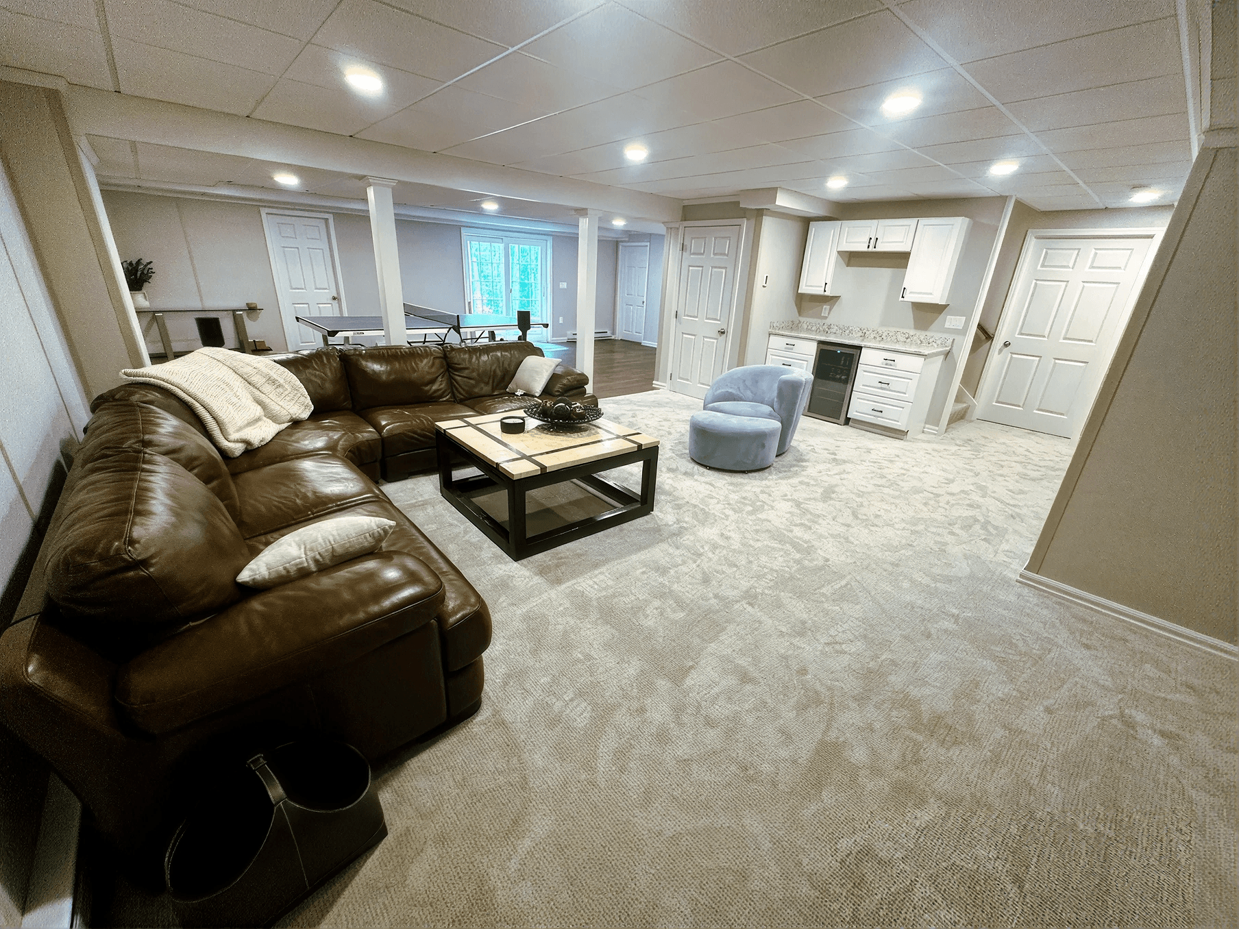 A beautifully finished basement designed as a spacious entertainment and family room featuring a large brown leather sectional sofa, a square coffee table with decorative accents, and a plush light gray area rug. In the background, a ping pong table sits in an open recreation area with large windows providing natural light, while a built-in wet bar with white cabinetry, granite countertops, and a beverage refrigerator offers convenient entertaining space. Recessed ceiling lights, neutral gray walls, white support columns, and soft carpeting create a bright, inviting atmosphere ideal for relaxing, hosting guests, or spending time with family.