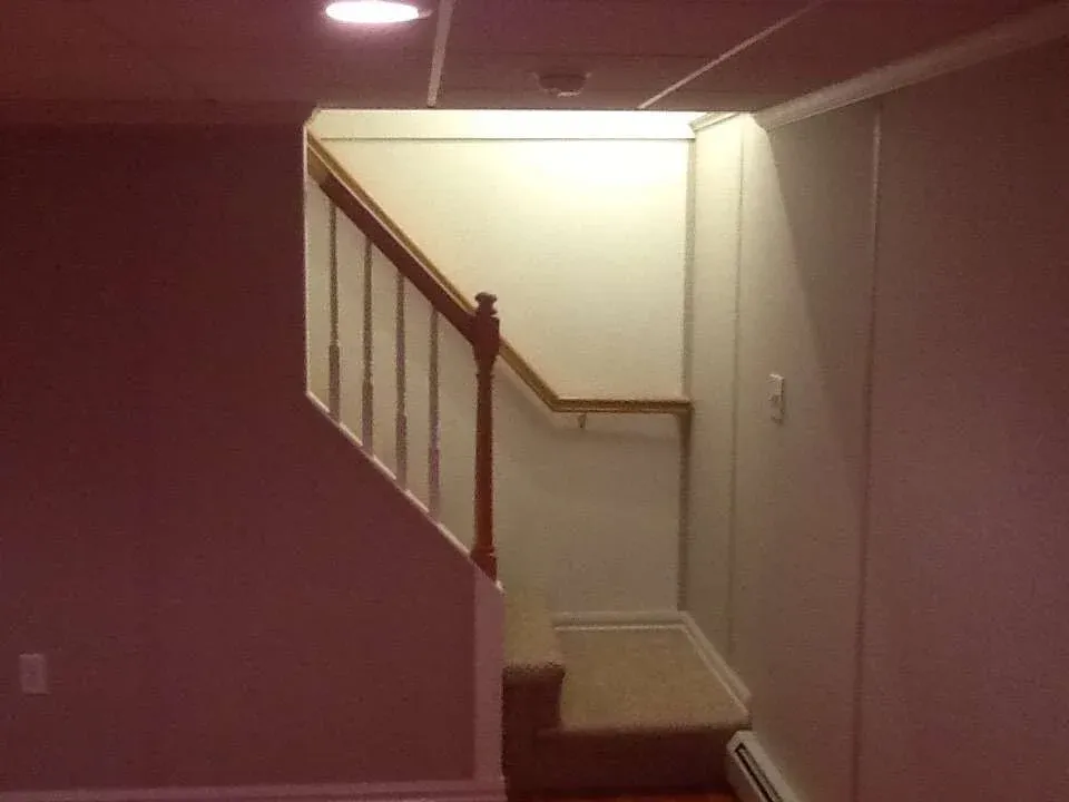 A dimly lit, small staircase with light-colored carpet leads upward, enclosed by beige walls. Minimalist design evokes a quiet, cozy ambiance.