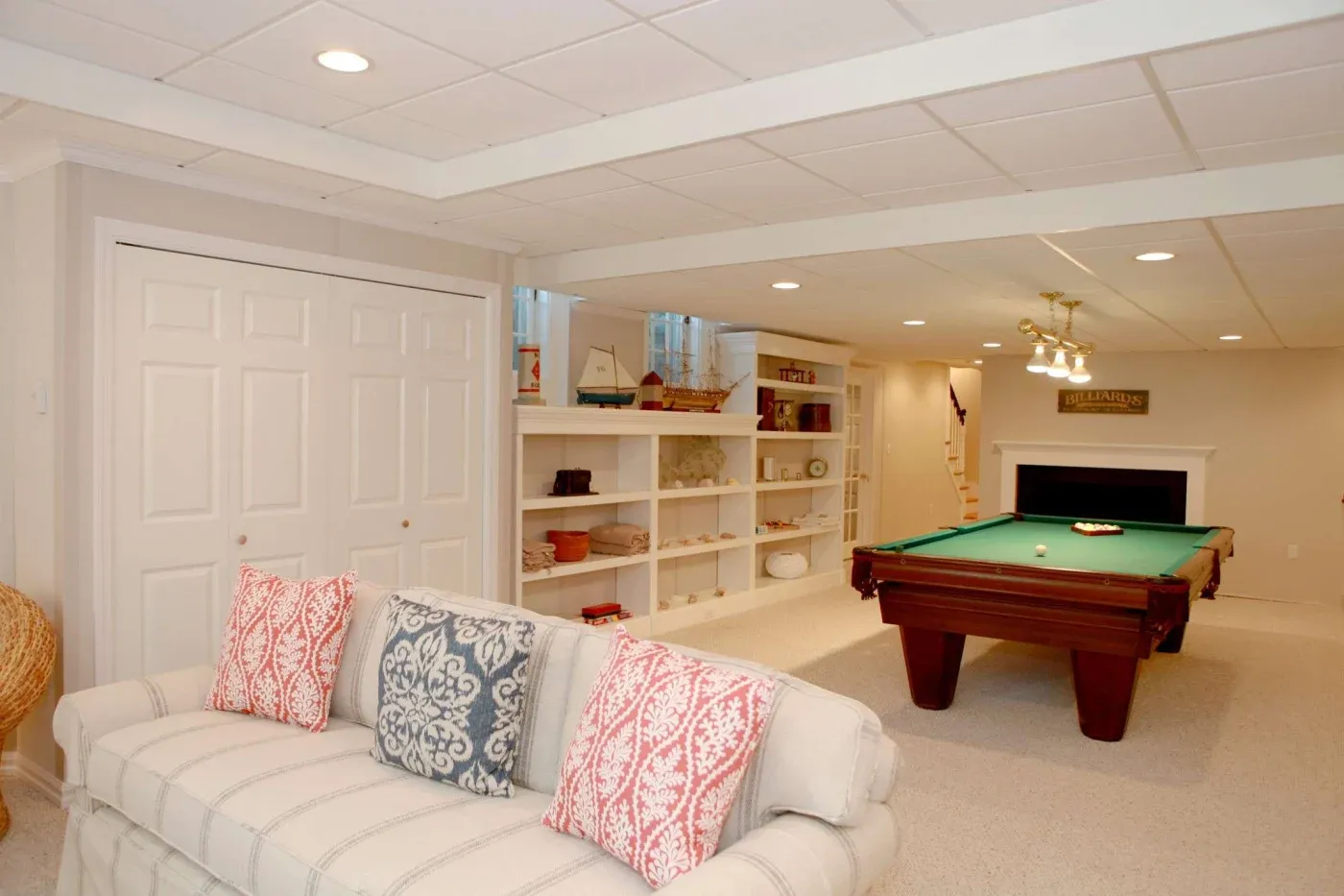 Stylish basement with a beige couch adorned with patterned pillows, facing a pool table. The room features built-in shelves and recessed lighting, creating a cozy, inviting atmosphere.