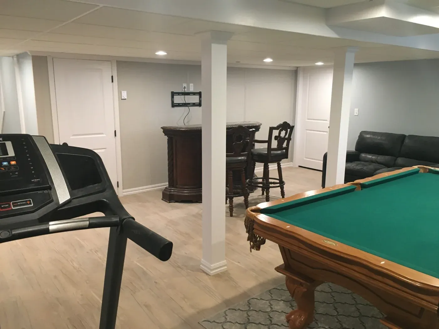 A cozy basement rec room with a treadmill, pool table with a green felt top, a small wooden bar with stools, and a black leather couch.