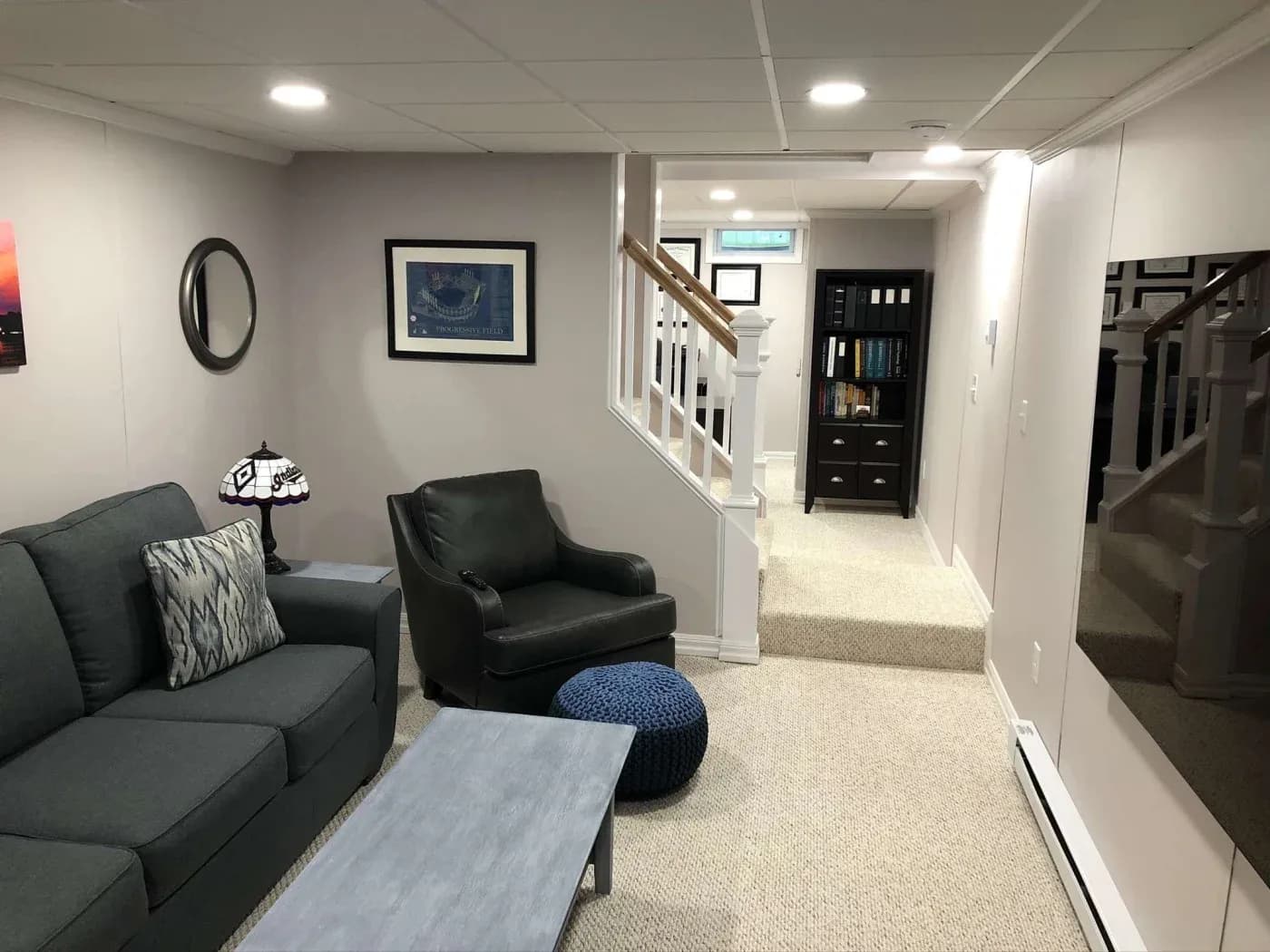 A cozy basement living room with a gray sofa, patterned cushions, an armchair, and a blue pouf. Framed art and a mirror adorn the walls, with soft lighting creating a warm ambience. A staircase and bookcase are visible in the background.