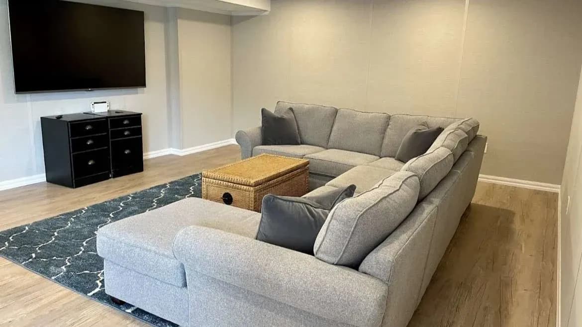 A cozy living room featuring a gray sectional sofa with dark cushions, a wicker coffee table, a black TV stand, and a large TV. The decor is modern and inviting.