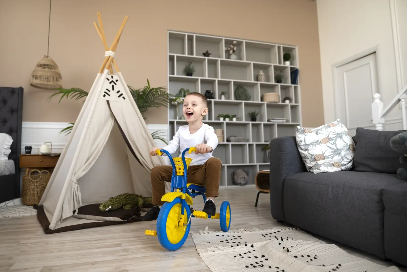 A joyful child rides a bright blue and yellow tricycle indoors, near a cozy teepee and a gray sofa, in a room with beige walls and shelves.