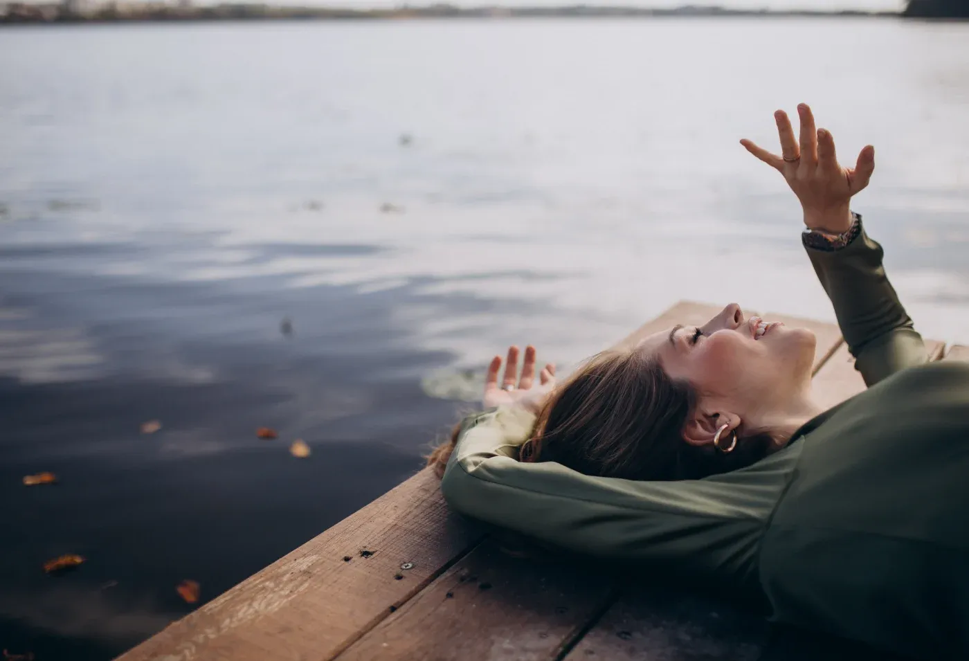 A woman in a green shirt lies on a wooden dock by a serene lake, gazing upwards. Her relaxed pose conveys tranquility and contemplation.