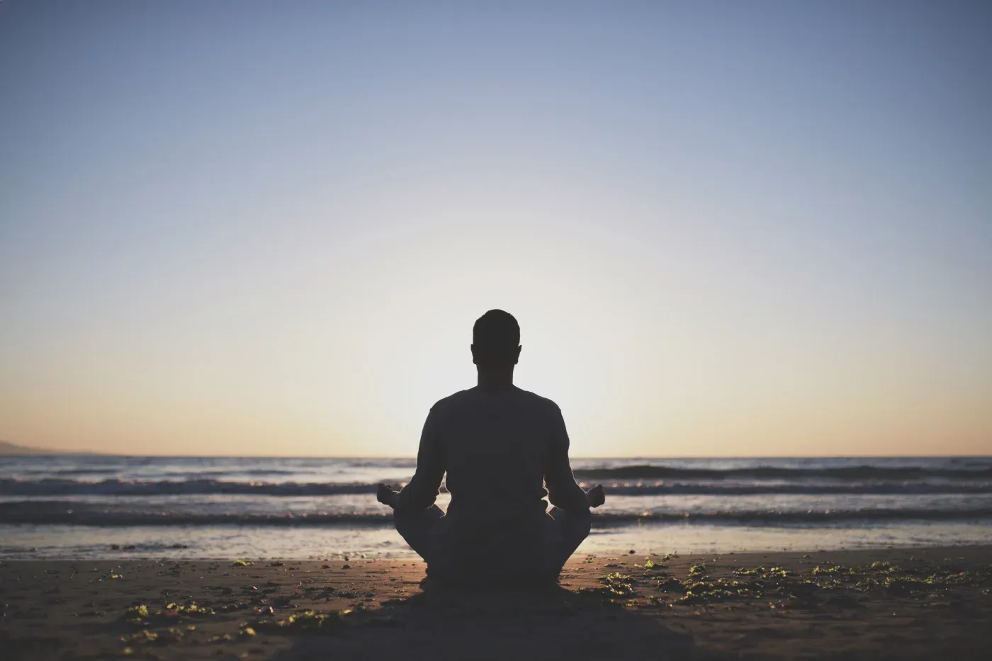 A person sits cross-legged on a beach at sunrise, facing the calm ocean. The sky is clear, creating a serene and peaceful atmosphere.