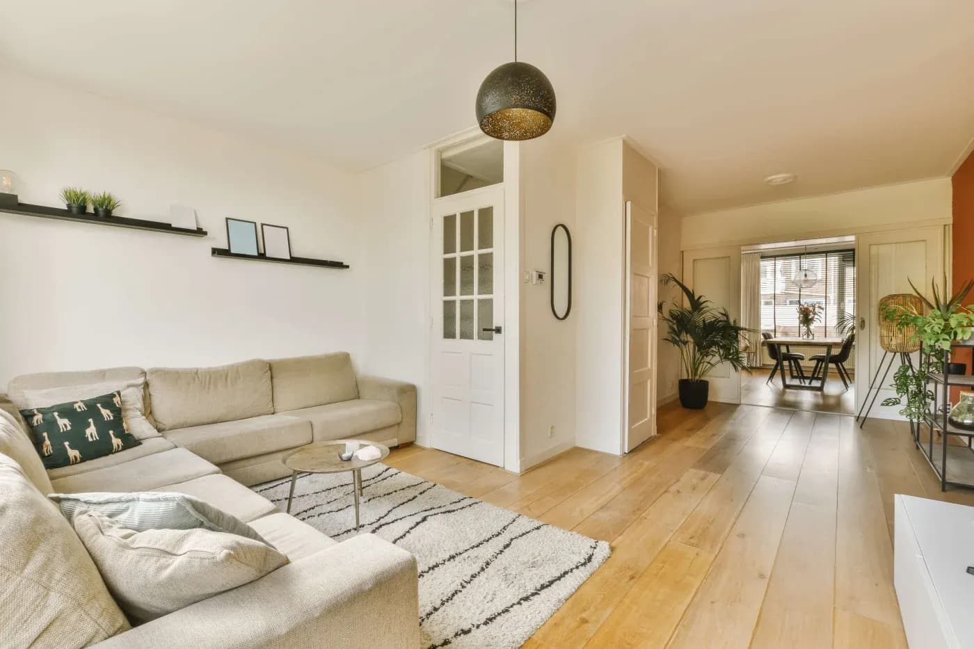 Bright and modern living room with beige L-shaped sofa, black-shelf decor, wooden floor, and a view into the dining area. Cozy, inviting atmosphere.