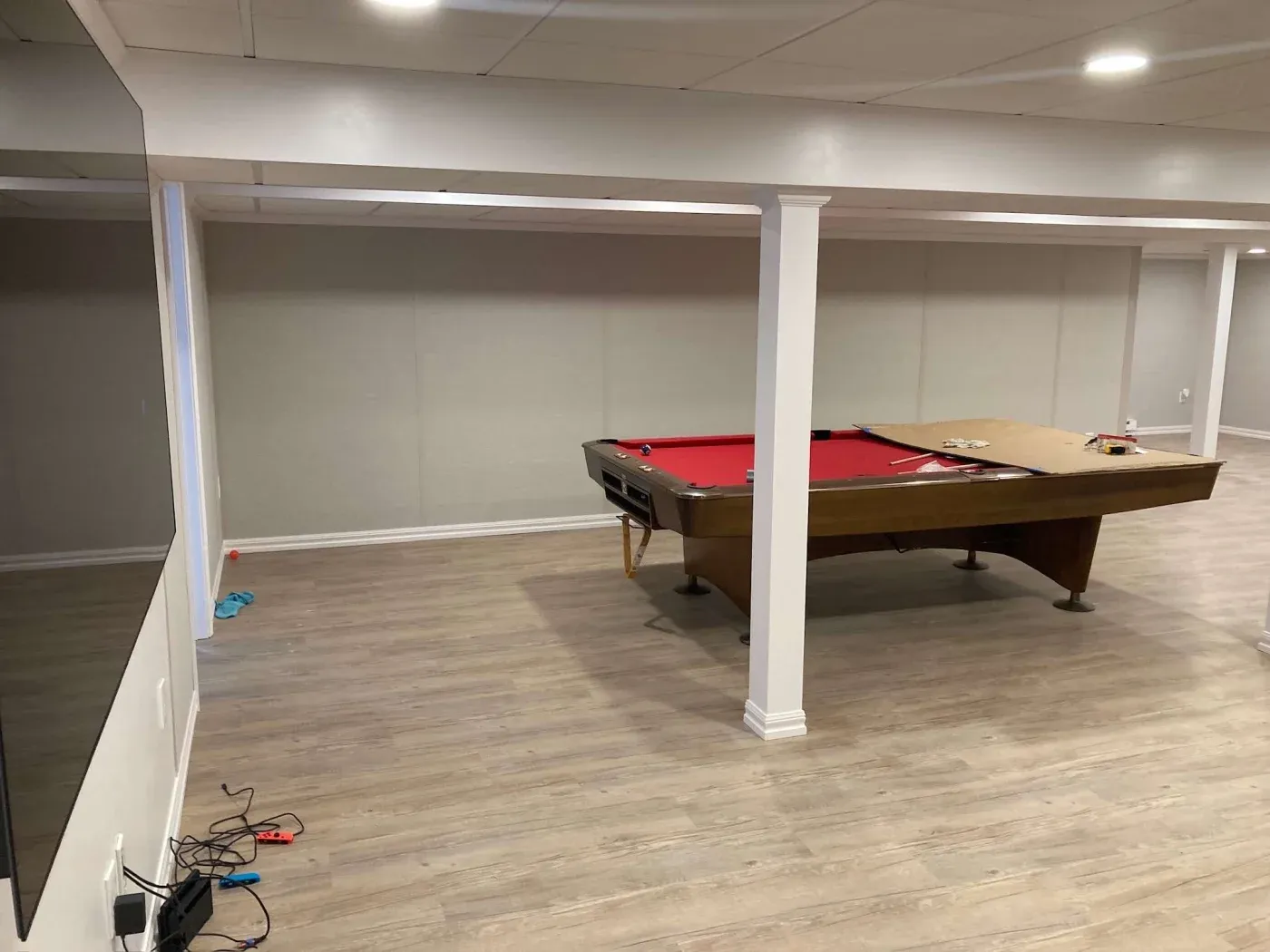 Spacious basement with wooden floors, red pool table under white ceiling beams, some tools and clutter on the surface, cables and items on the floor.