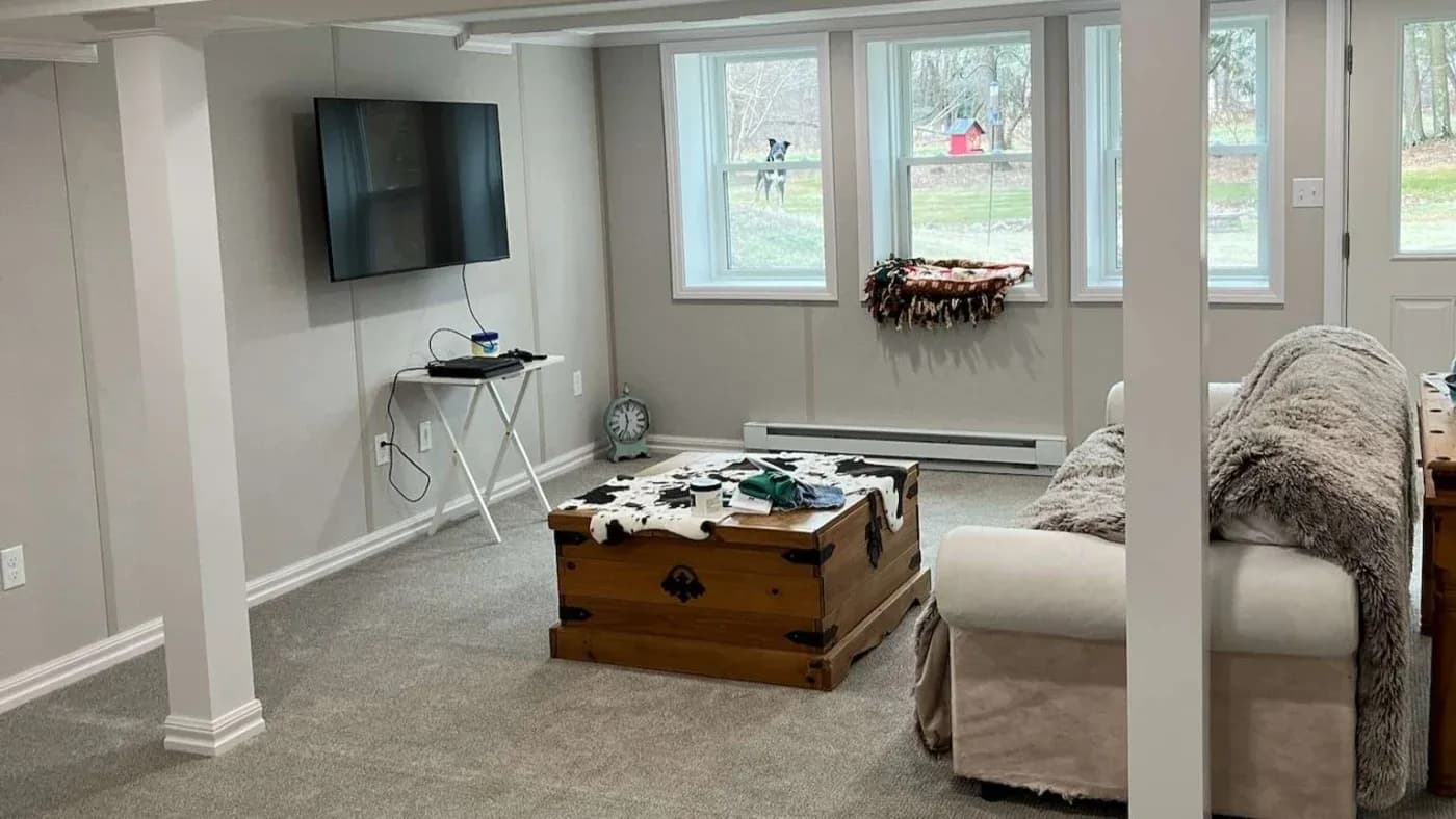 Cozy basement with a beige sofa, rustic wooden chest, and wall-mounted TV. Dog peeks through large windows, adding warmth to the bright space.