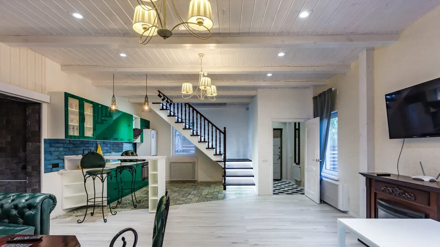 Spacious open-plan living area with green kitchen cabinets, wooden staircase, and vintage decor. Soft lighting creates a warm, inviting atmosphere.