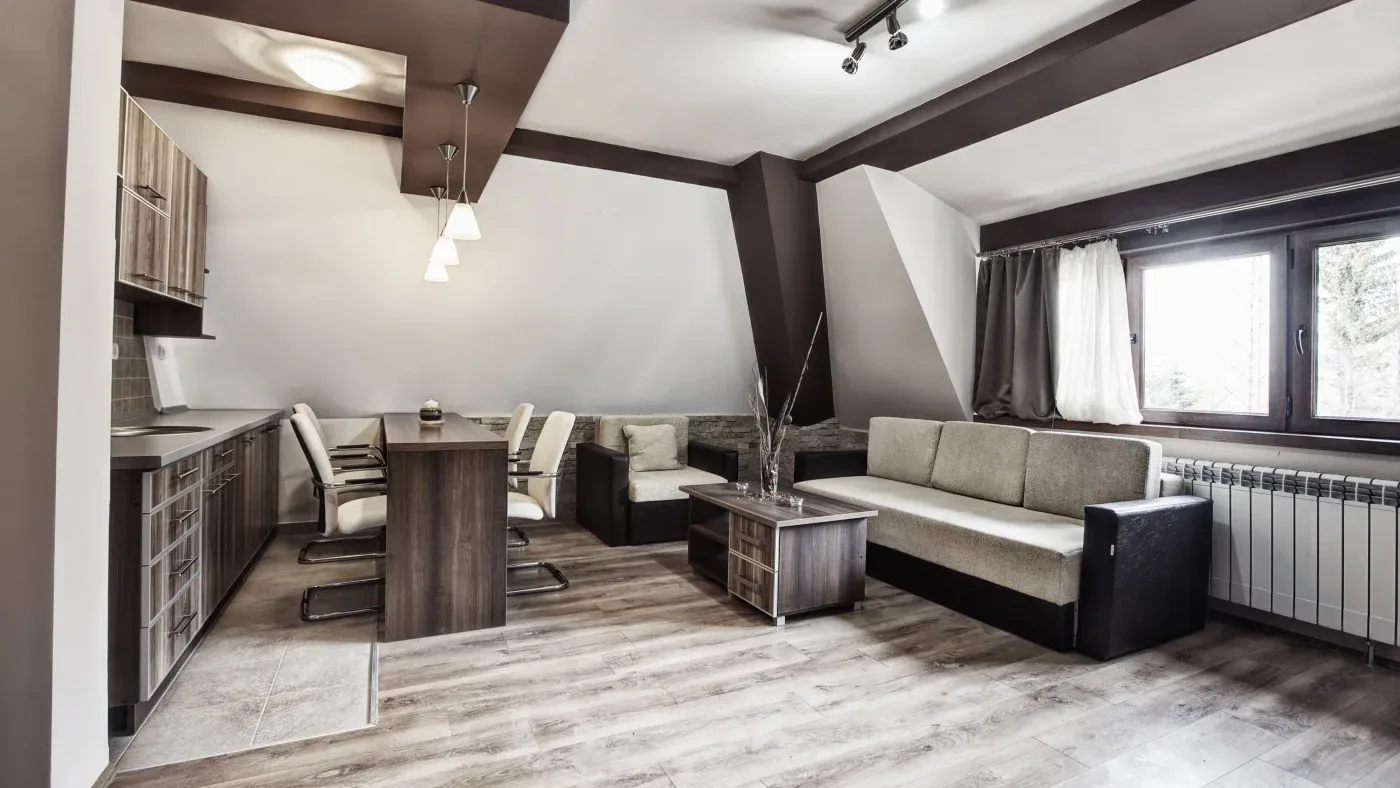 Modern studio apartment with wooden floors and brown accents. Features include a compact kitchen, dining table, cozy seating area, and large window.