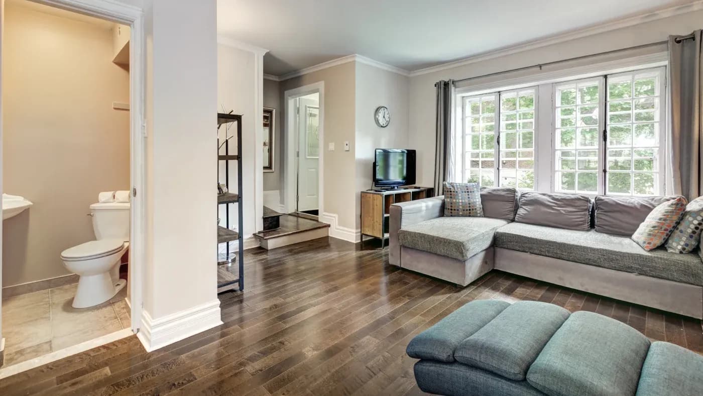 Bright living room with hardwood floors, featuring a gray sectional sofa, a TV on a wooden stand, large windows with curtains, and a view into a small bathroom.